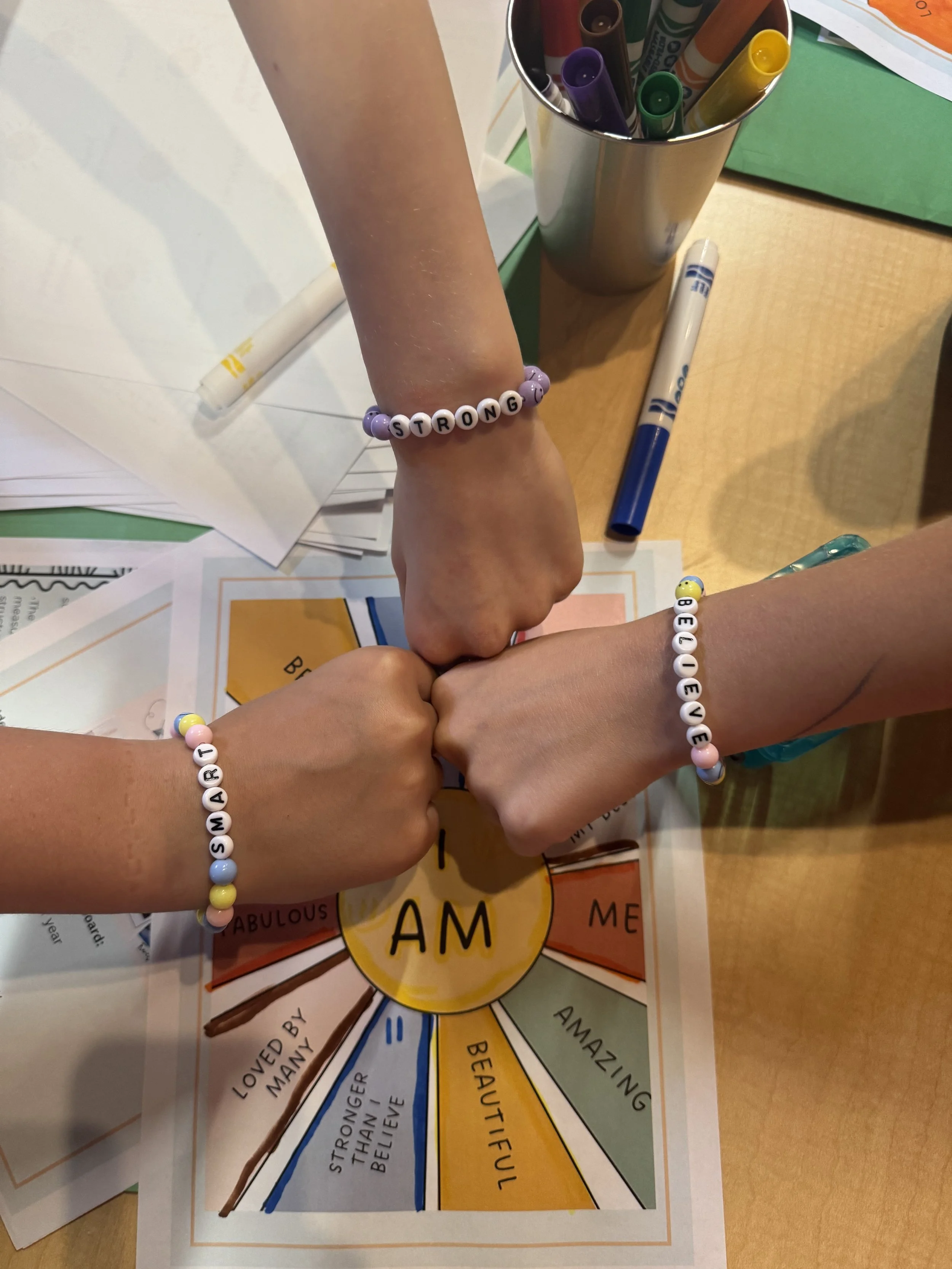Four hands wearing friendship bracelets with words like 'STRONG,' 'BELIEVE,' 'BEAUTIFUL,' and 'AMAZING,' joined together over a colorful infographic or worksheet on a desk, with markers and papers nearby.