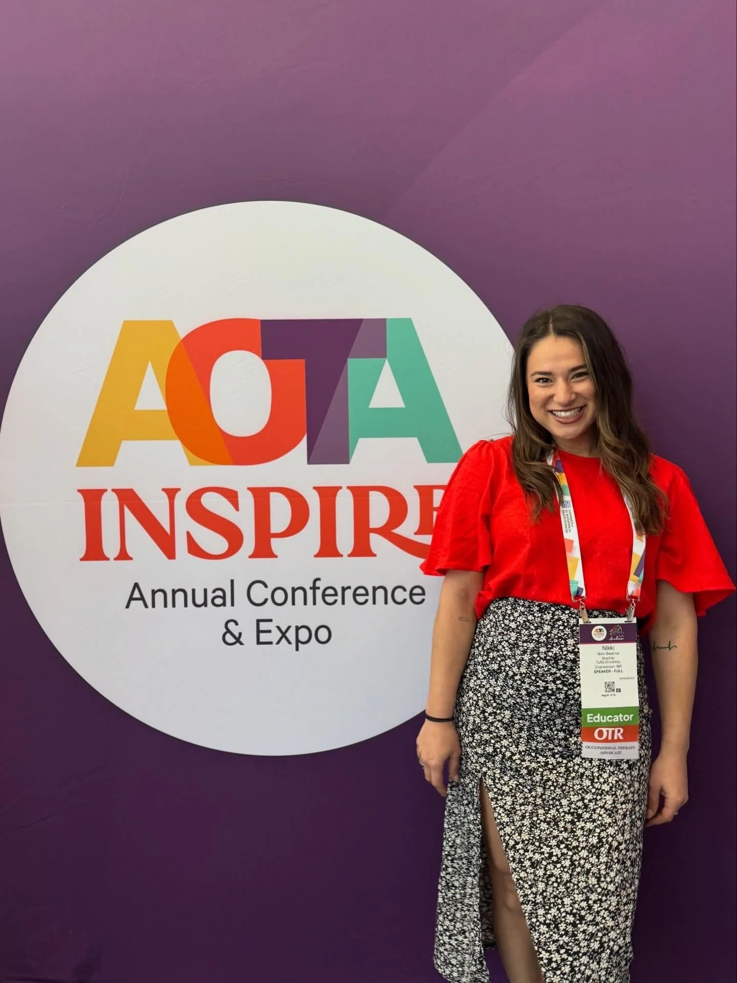 A woman smiling at an indoor conference and expo, standing in front of a purple wall with a large round sign that reads "AINSPIRE" and "Annual Conference & Expo." She is wearing a red top, a black and white patterned skirt with a high slit, and has conference lanyard and badge that says "Educator."