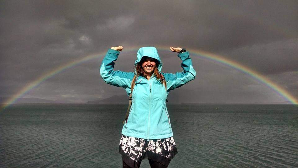 Woman in a turquoise rain jacket and black and white patterned skirt standing in front of ocean with rainbow and dark cloudy sky in background.