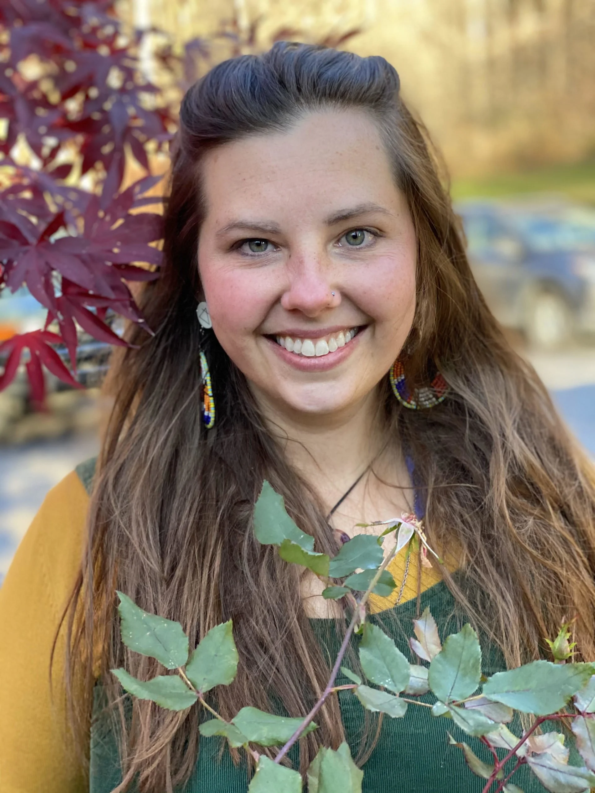 Close-up of a smiling woman with long brown hair, blue eyes, freckles, and a nose piercing. She is outdoors with fall foliage in the background, holding green leaves and branches.
