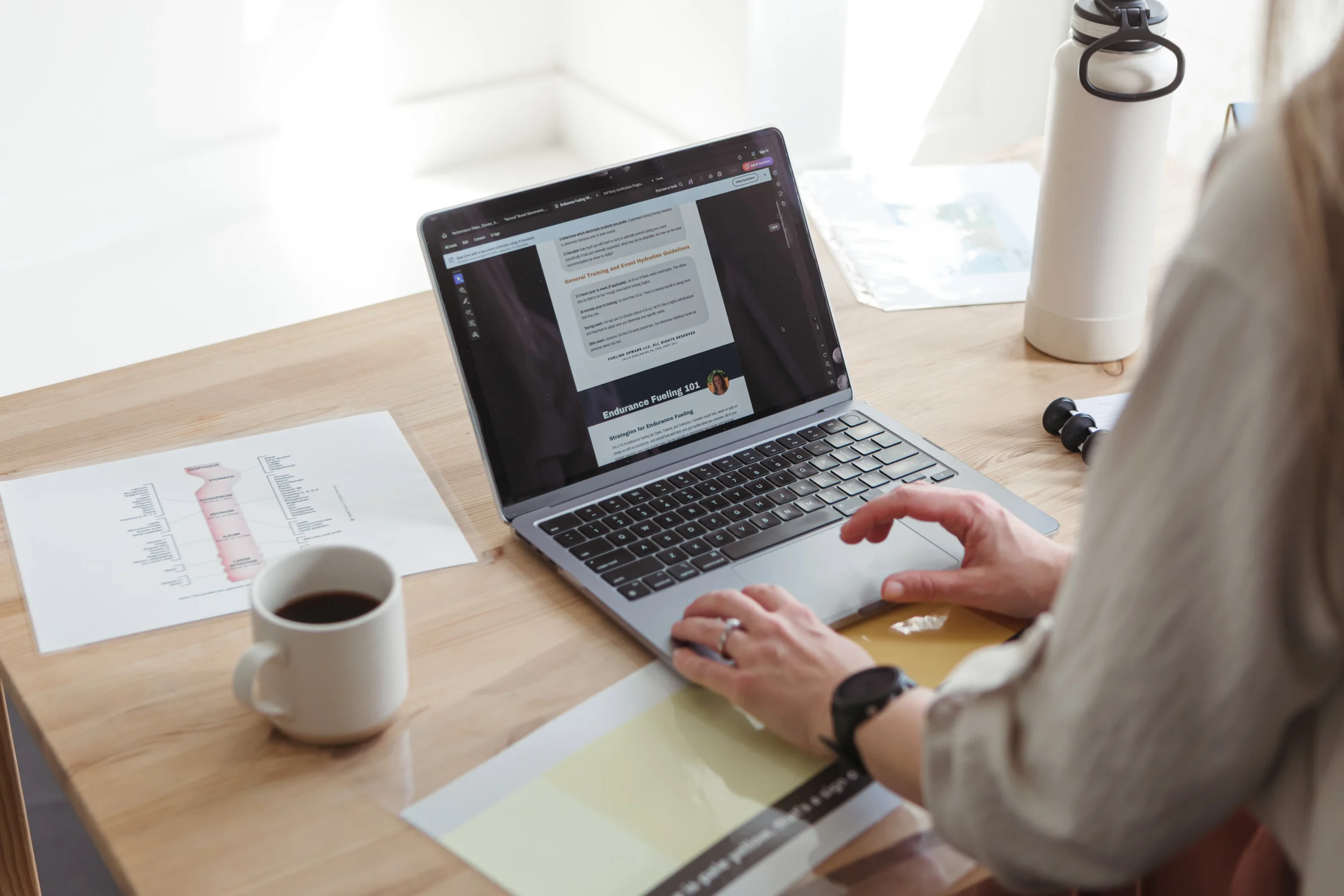 A person working on a laptop at a desk, with a coffee mug in front, a water bottle nearby, and printed documents on the table.