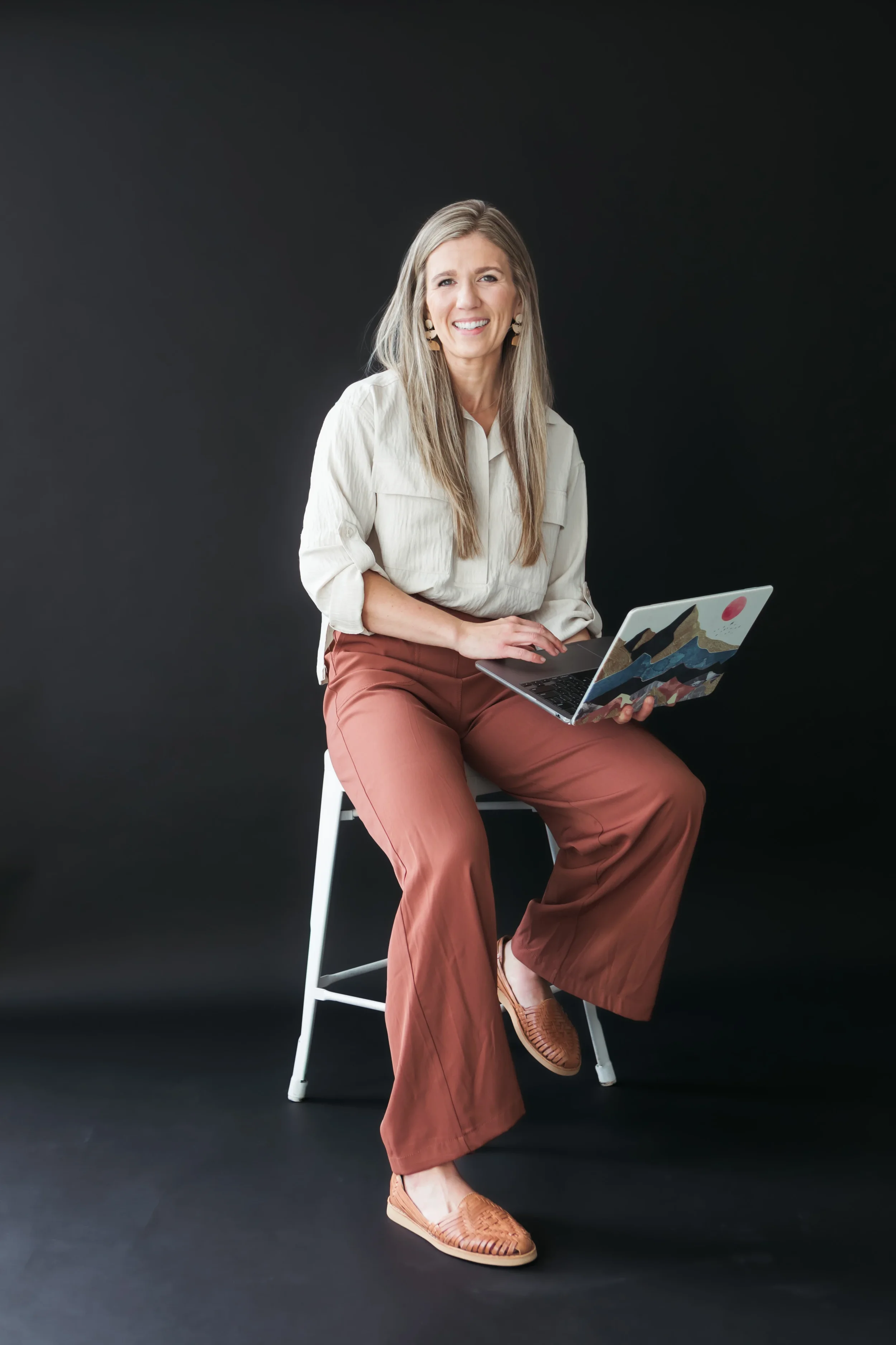 A woman with long blonde hair smiling, sitting on a white chair against a black background, holding a laptop with a mountain landscape sticker, wearing a cream blouse, rust-colored pants, and tan woven shoes.