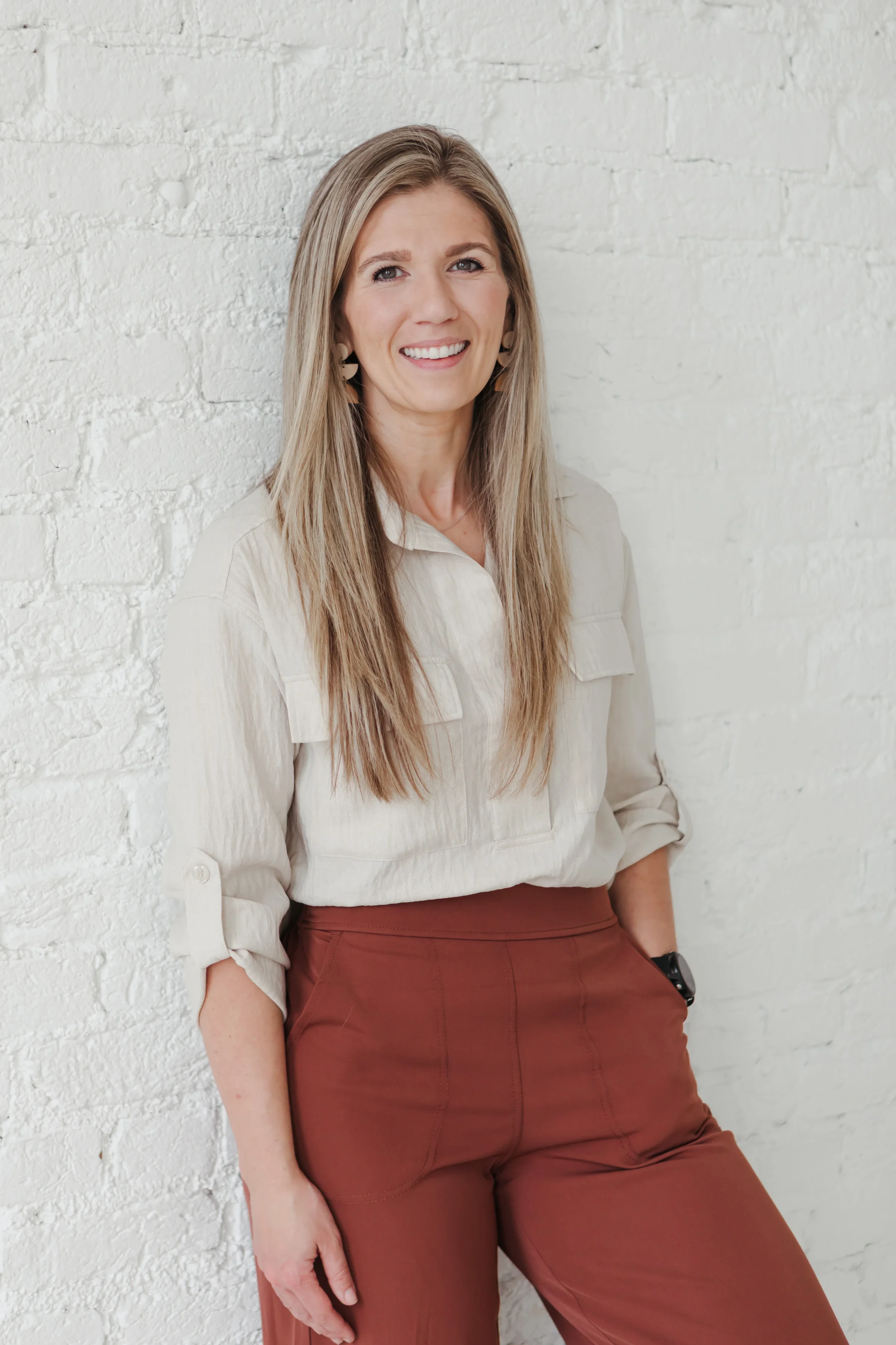 A woman with long blonde hair, wearing a beige shirt with rolled sleeves and rust-colored pants, standing against a white brick wall, smiling at the camera.
