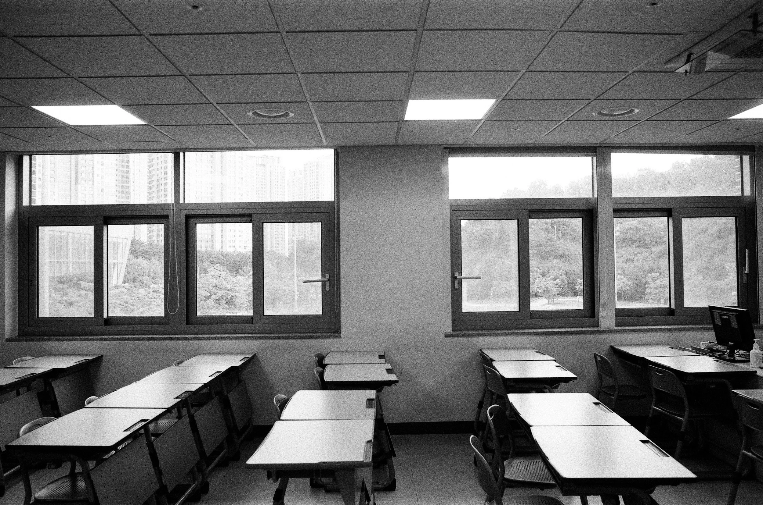 An empty classroom with desks and chairs arranged in rows, large windows at the back showing a cityscape and trees, and a tiled ceiling with fluorescent lights.
