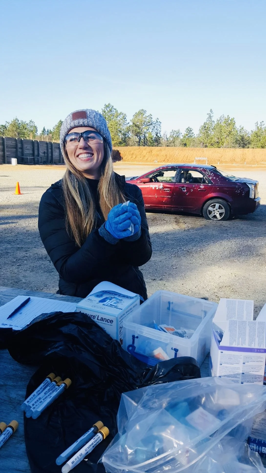 A woman with long blonde hair smiling, standing outdoors on a shooting range beside a car with dents, medical supplies on a table, and orange traffic cones in the background, wearing a cream beanie, glasses, black coat, and medical gloves.