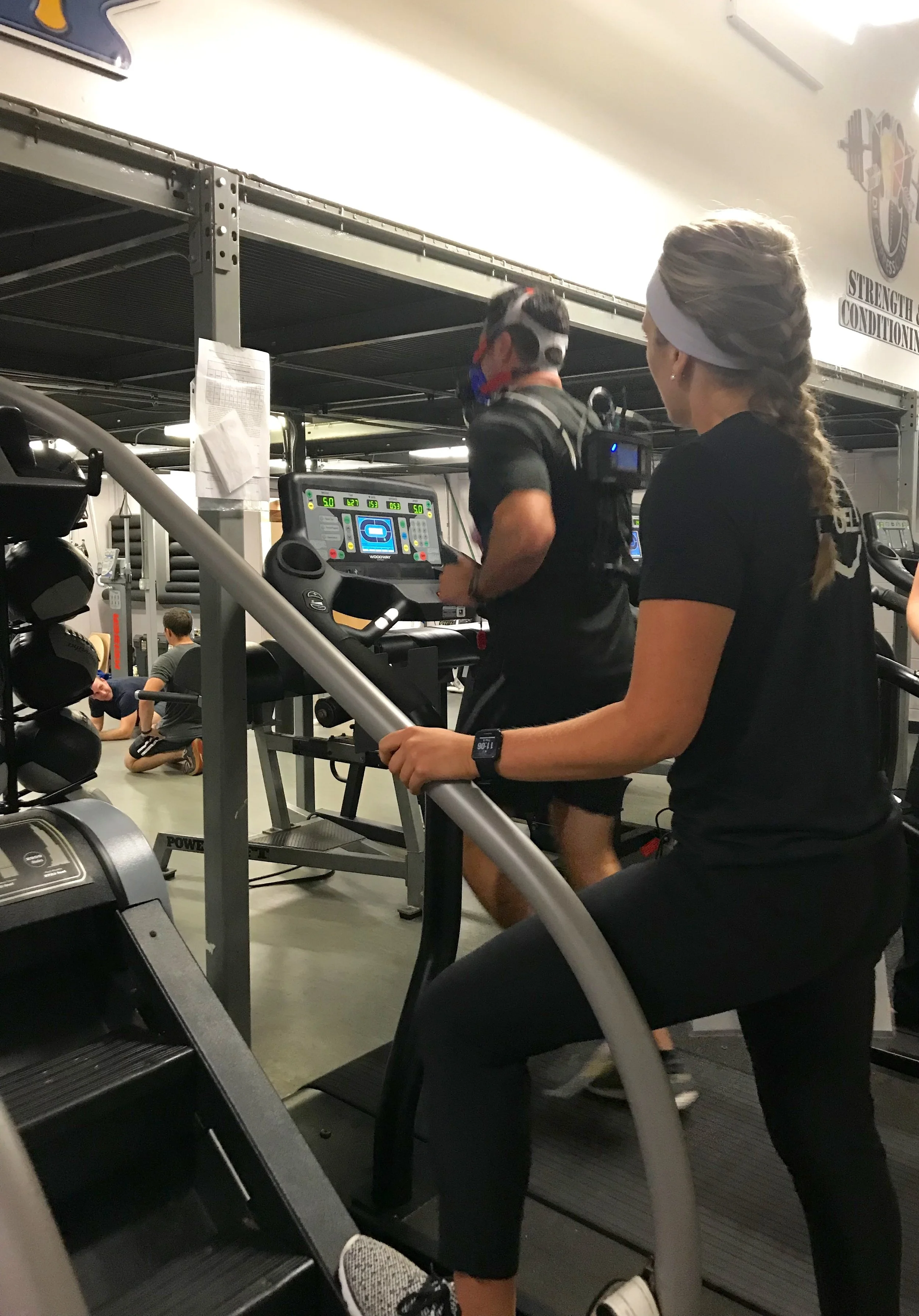 A woman and a man are on treadmills in a gym. The woman is wearing a black shirt, black pants, sneakers, and a white headband. The man is wearing black workout clothes, a mask, and is running on a treadmill with digital display. There are other gym members in the background, and gym equipment is visible.
