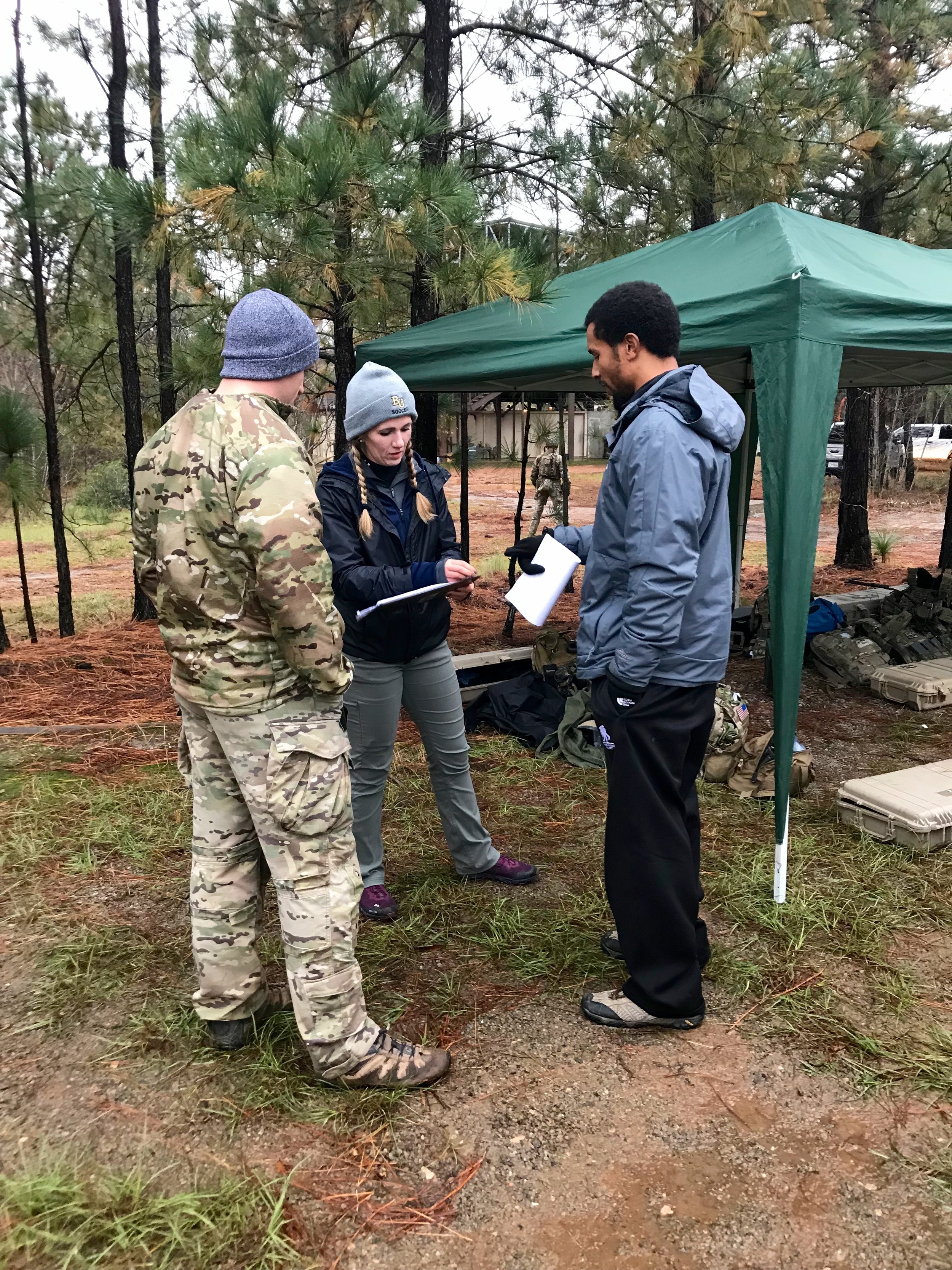 Three people are engaged in conversation outdoors, standing under a green canopy tent in a forested area with pine trees. One man is dressed in camouflage, a woman in a navy jacket and green pants with a beanie, a man in a gray jacket and black pants