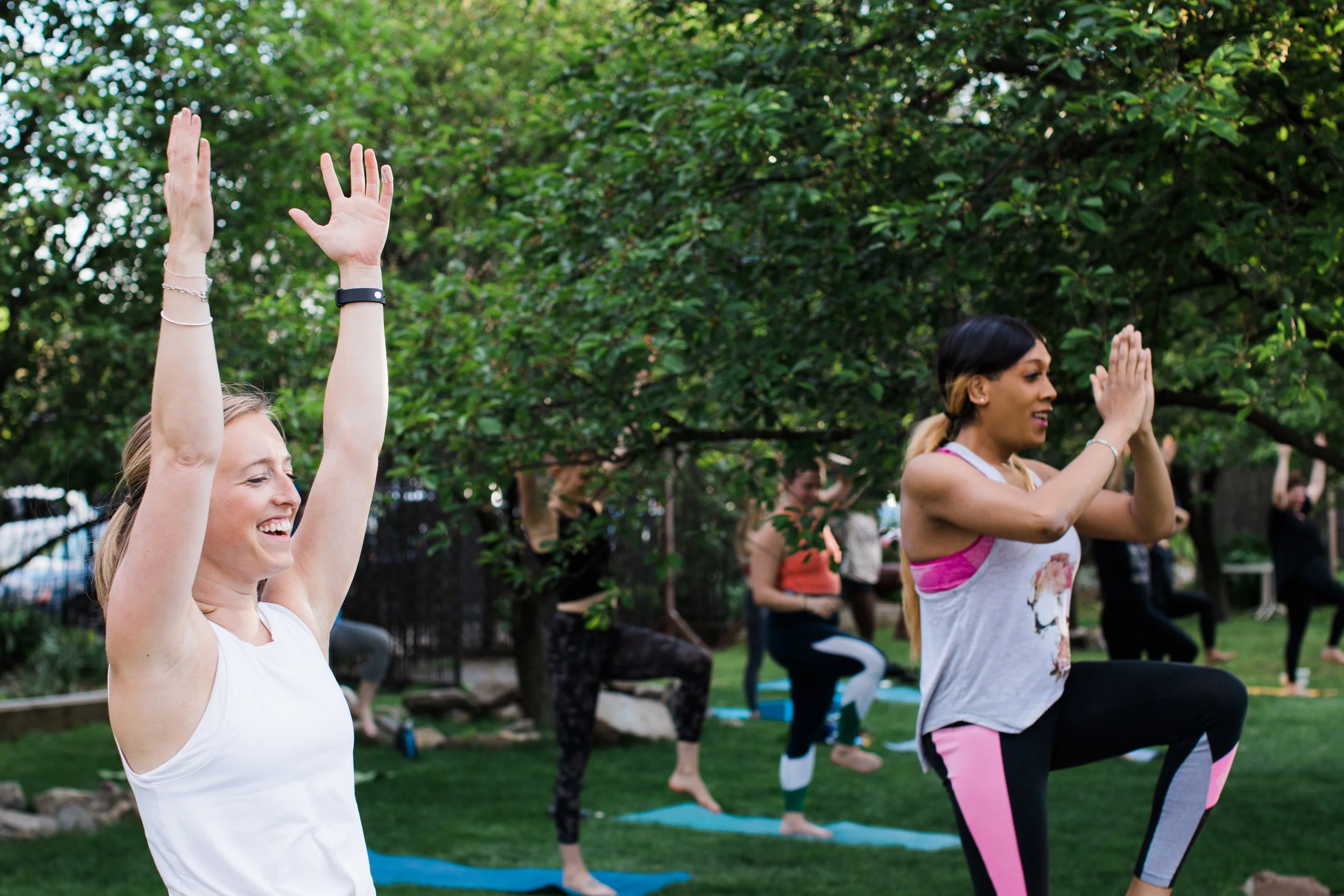 Women participating in outdoor yoga or exercise class, standing on yoga mats in a park with green trees, raising their arms.