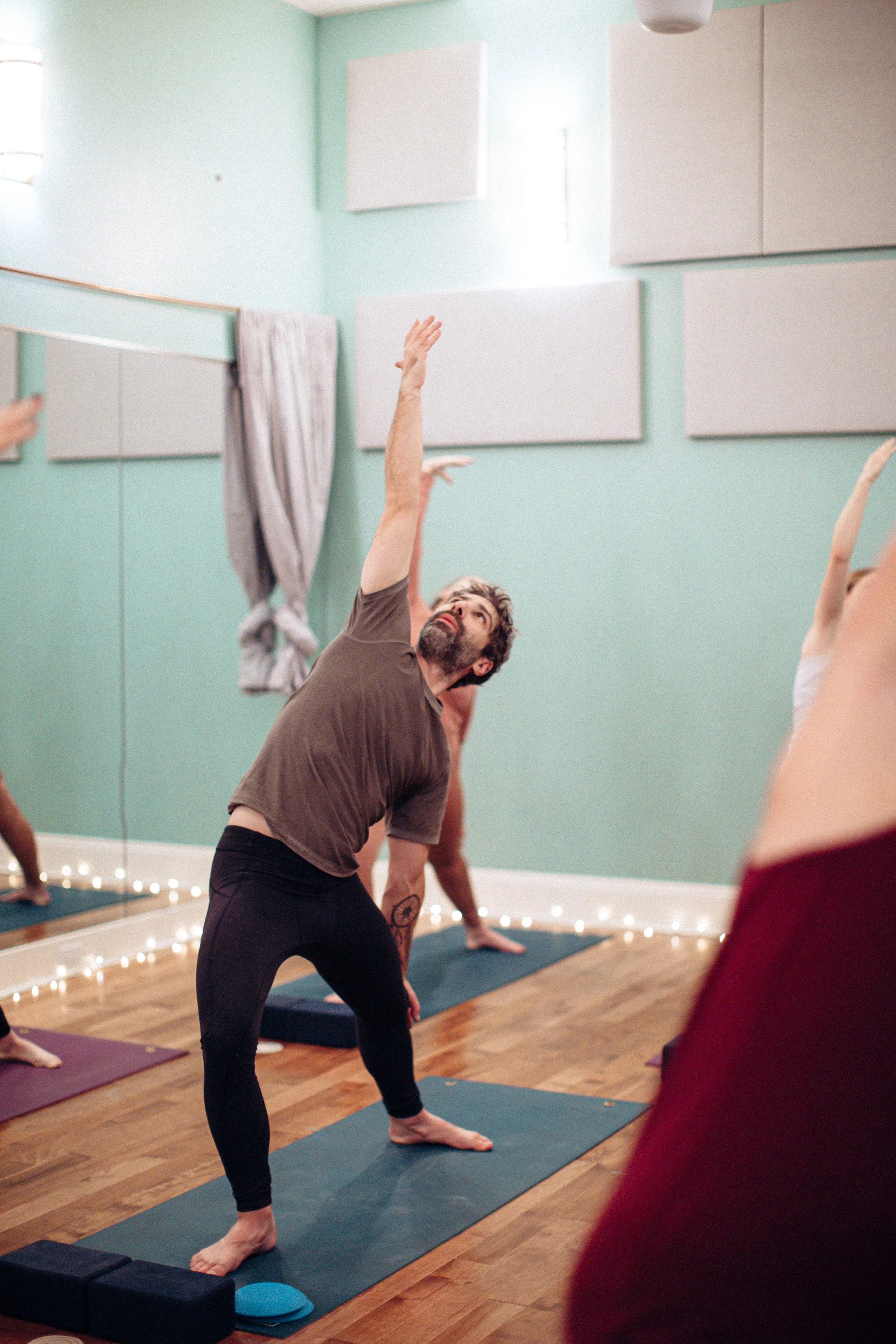 A man with dark hair and beard practicing yoga in a studio with light green walls, wooden floor, and acoustic panels, surrounded by yoga mats and blocks.