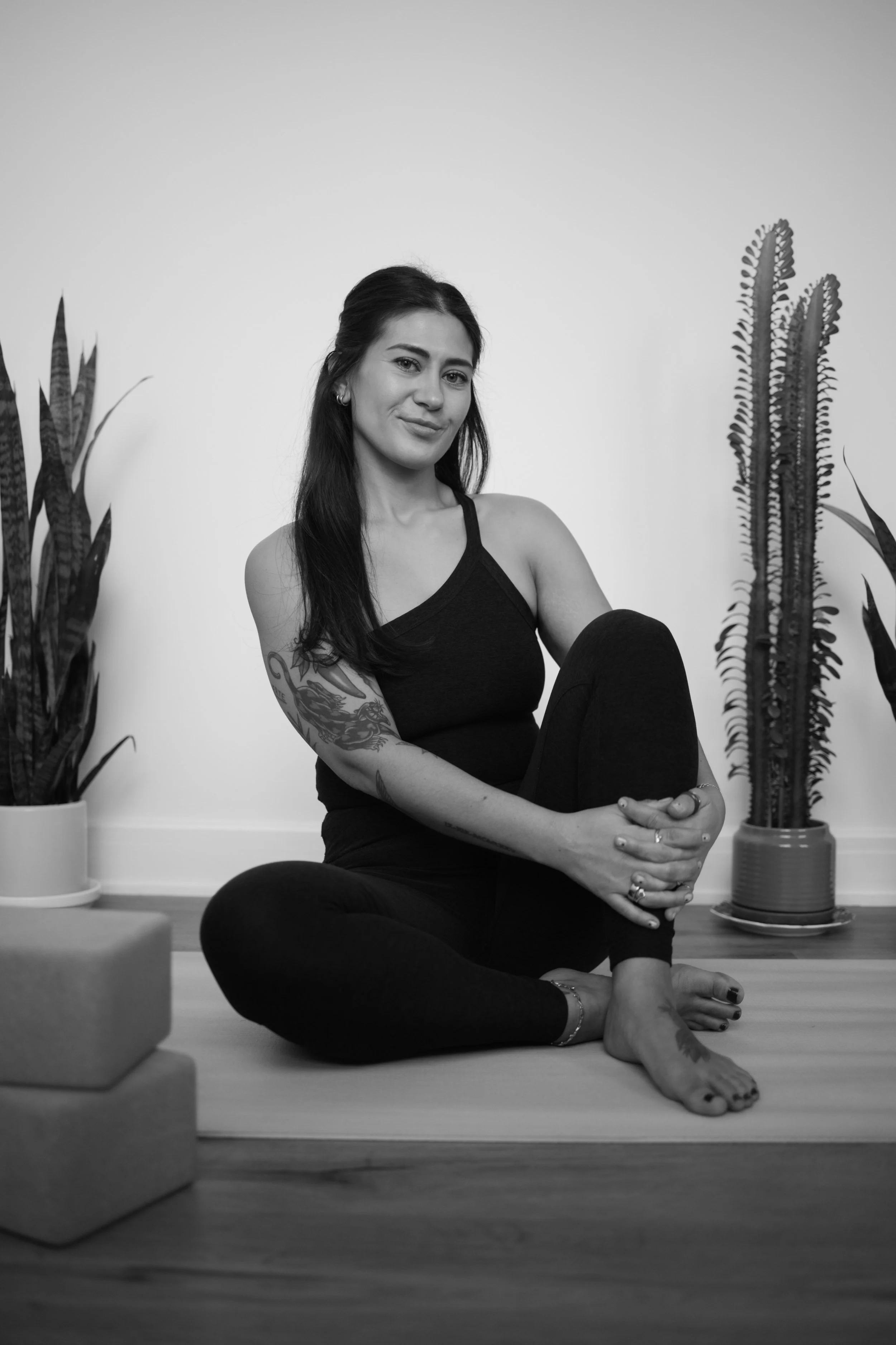 A woman practicing yoga indoors, sitting cross-legged on a yoga mat, surrounded by potted plants, with a serene expression.