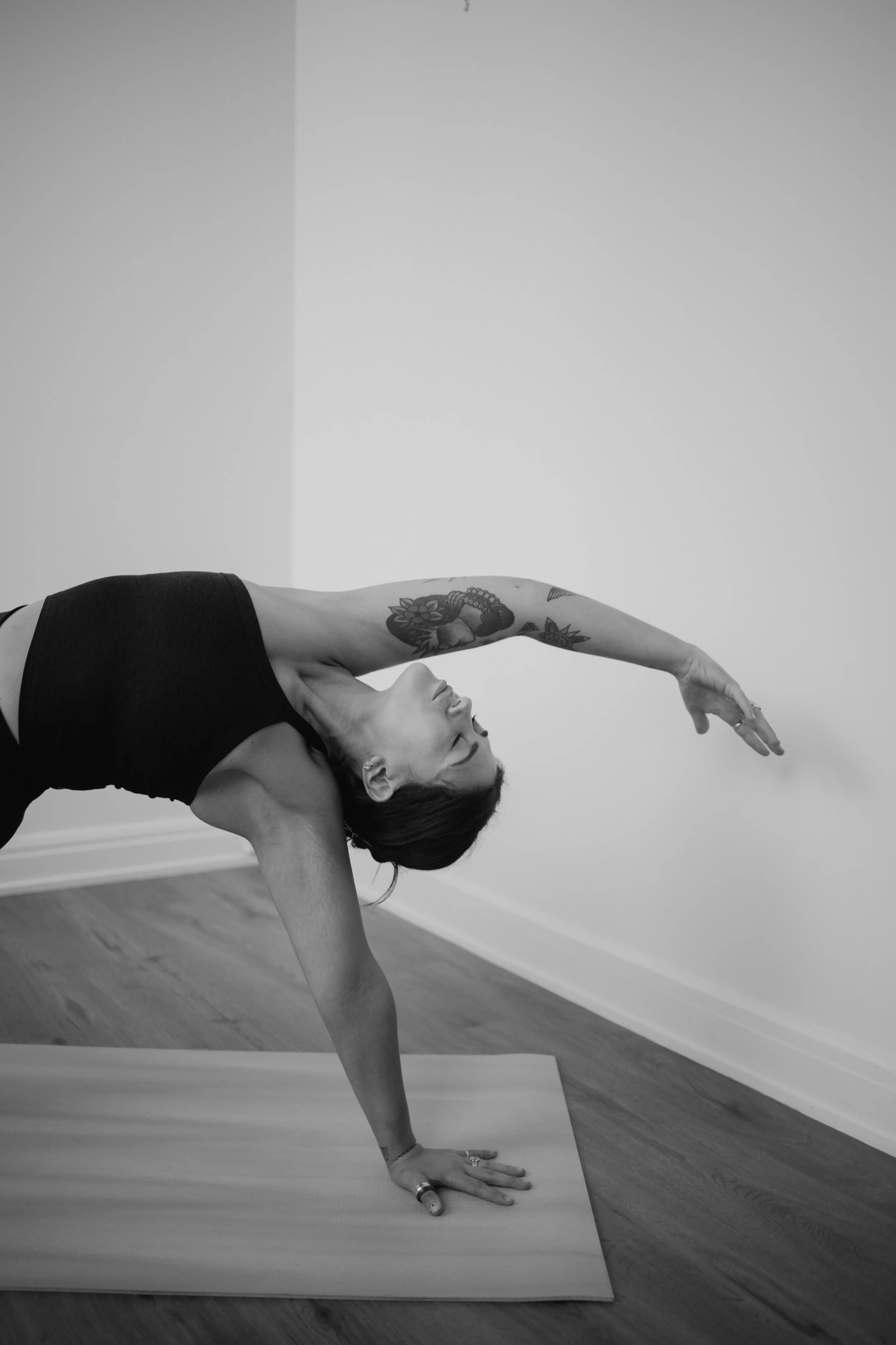 A woman practicing yoga indoors, performing a side plank pose with one arm extended overhead, wearing a black sports bra and tattooed arm, on a yoga mat in front of a plain wall.
