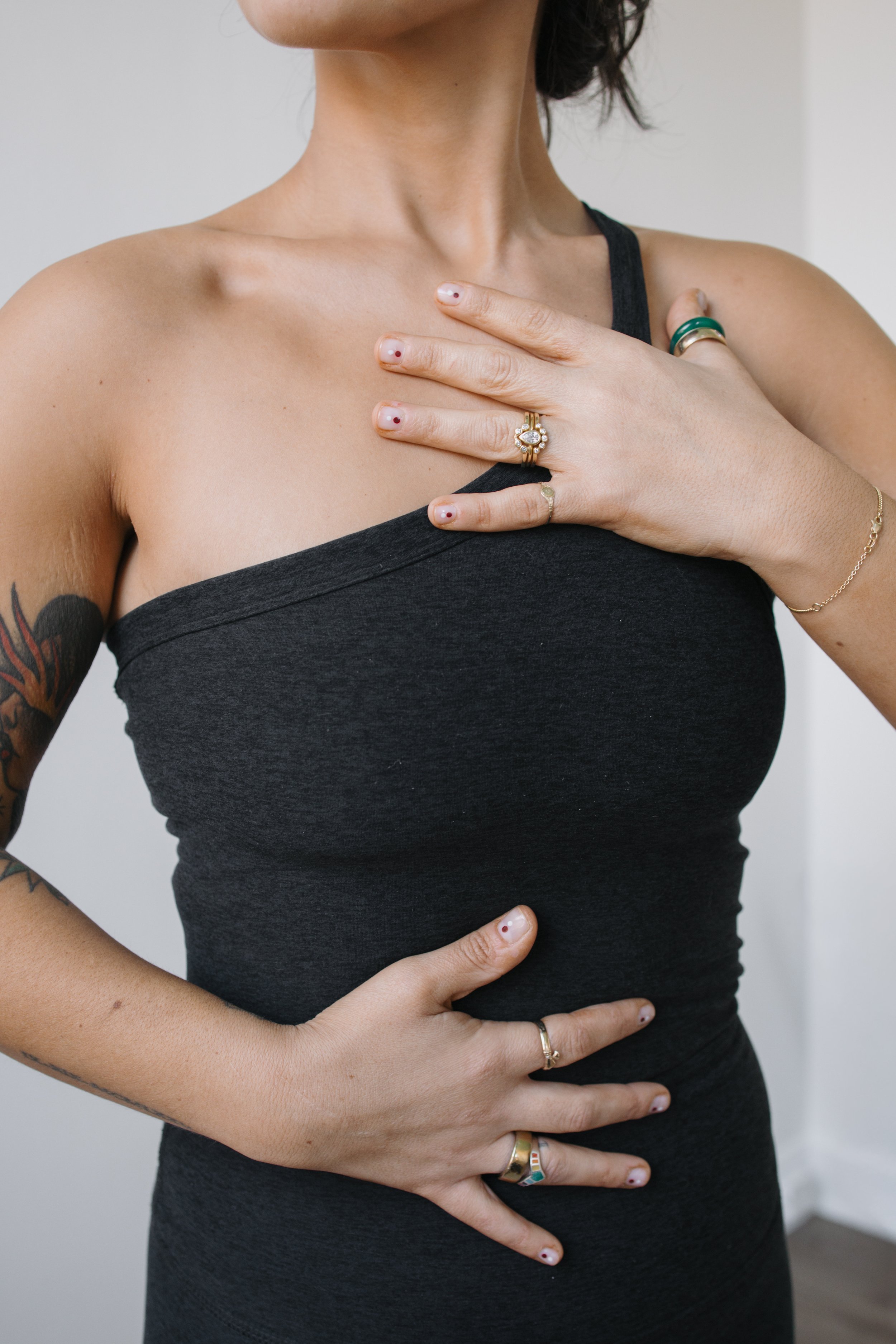 Close-up of a person wearing a black tank top, with their left hand placed on their chest and right hand on their stomach, showing rings, bracelets, and tattoos.
