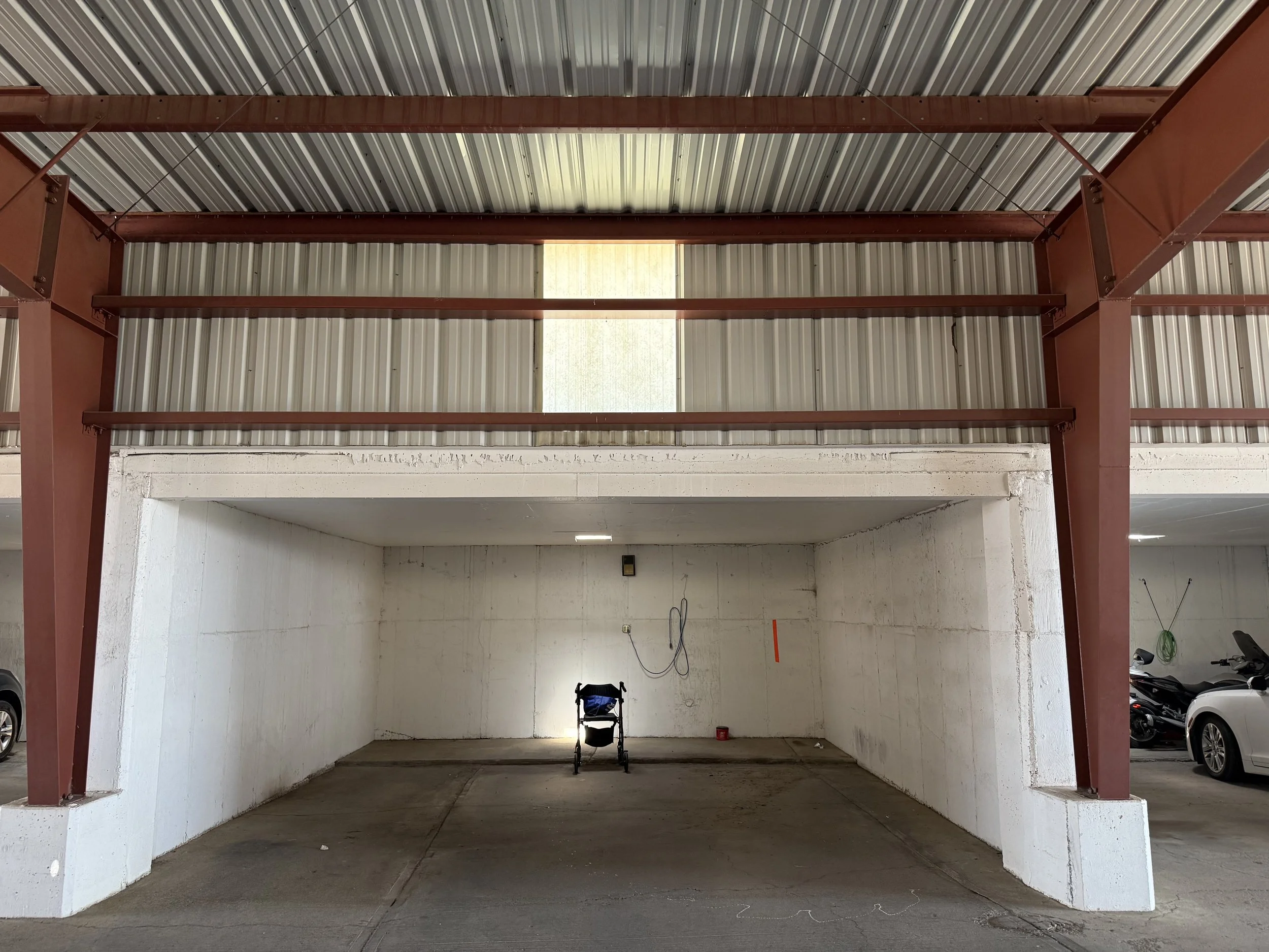 An indoor parking garage with a large empty space, white concrete walls, the ceiling has metal roofing panels, and a small window letting in natural light.