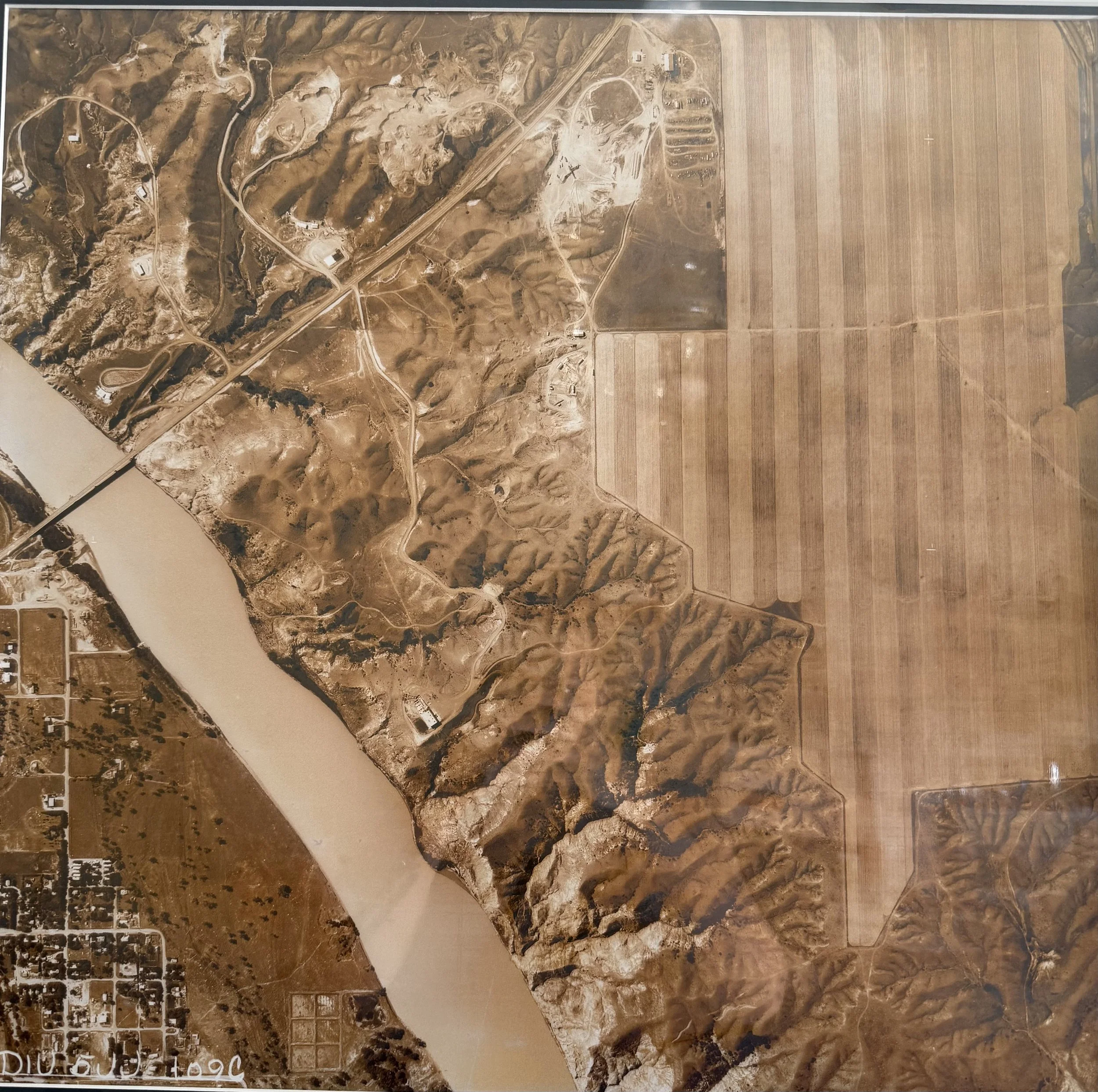 Aerial view of a landscape showing a river on the left, surrounding rugged hills and mountains, and flat agricultural fields on the right.