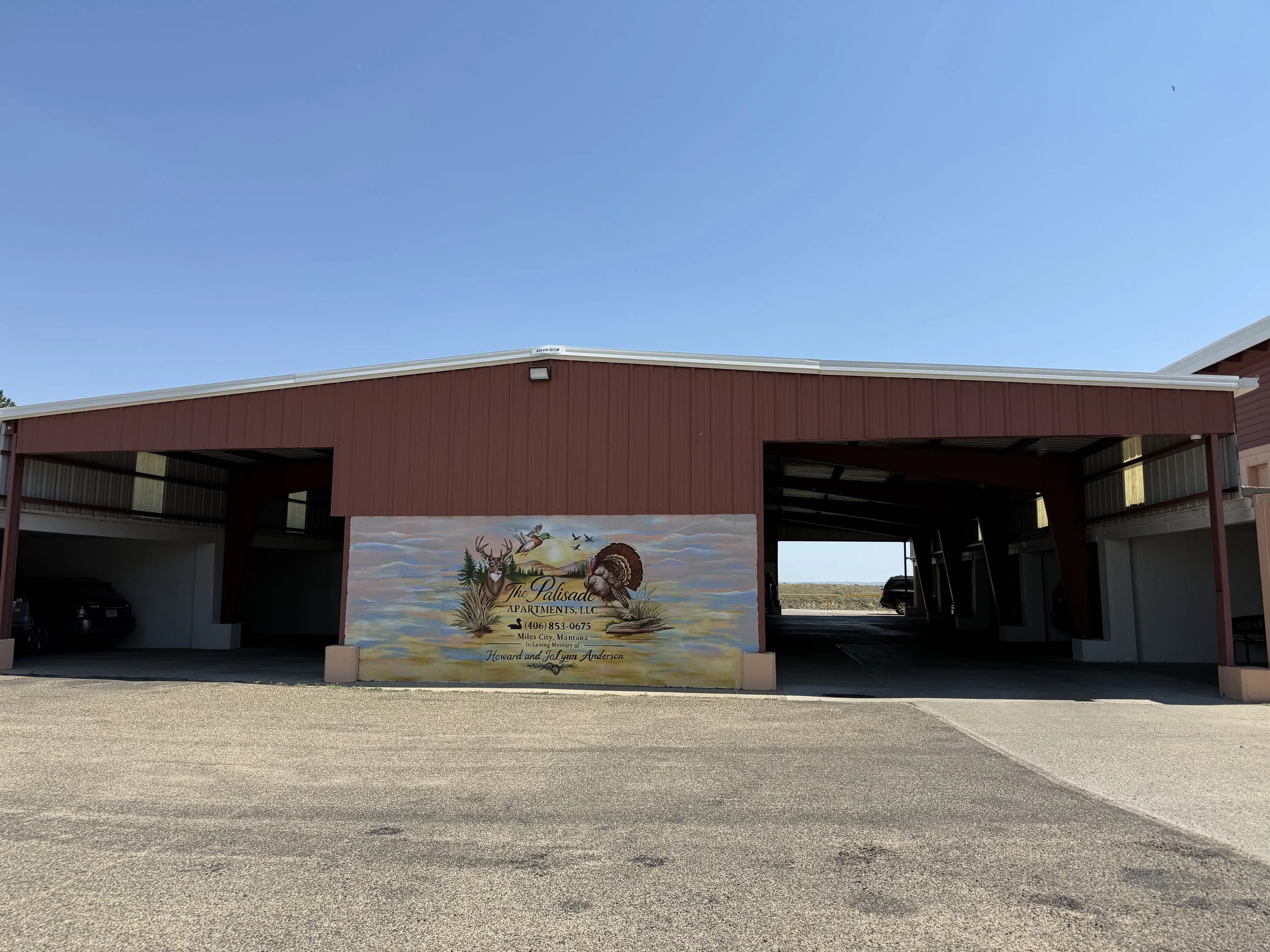 A red barn-style building with an open front and a large painted mural on the side. The mural depicts deer, trees, a flying eagle, and a sunset or sunrise sky. There is a sign on the building that reads "The Palisade Apartments, LLC," along with a phone number and the names Howard and Jo Lynn Anderson. The sky above is clear and blue, and the parking lot in front of the building is empty.