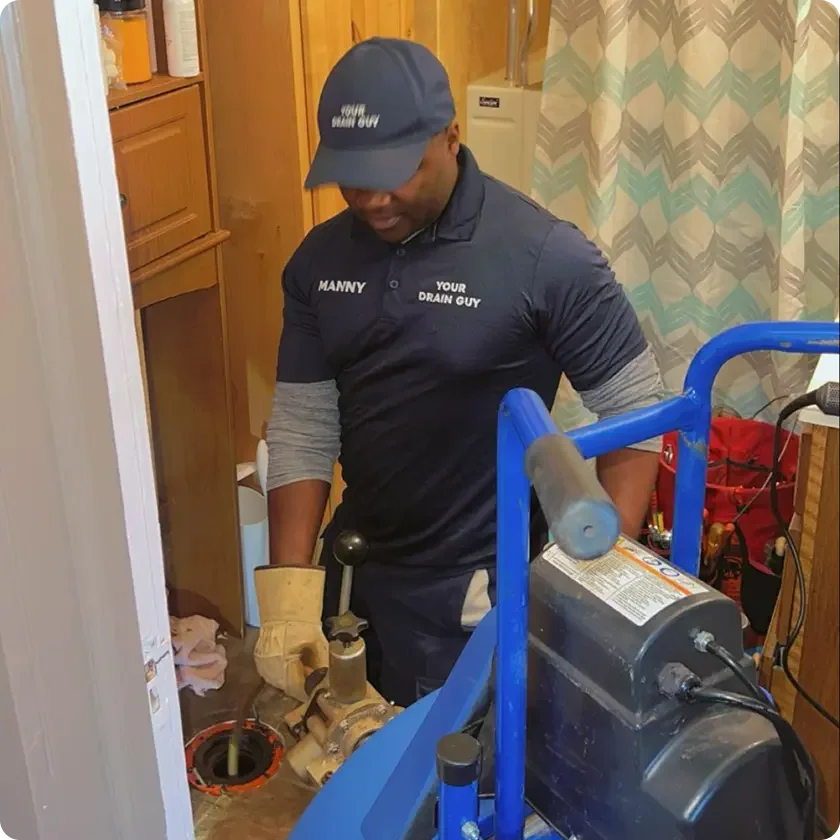 Man wearing a dark uniform and cap working on a plumbing pipe with tools inside a kitchen or utility room.
