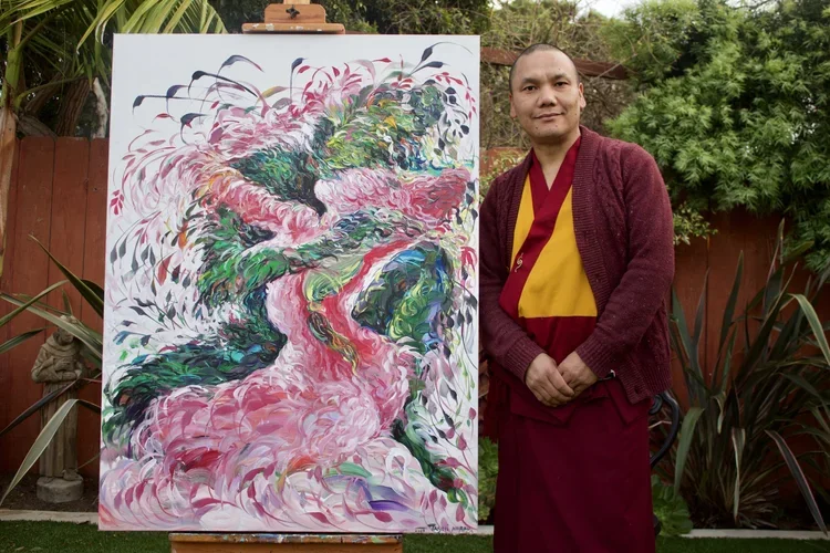 A man dressed in maroon and yellow traditional clothing standing next to an abstract painting of a pink dragon with green, blue, and purple accents, outdoors with greenery and a wooden fence in the background.