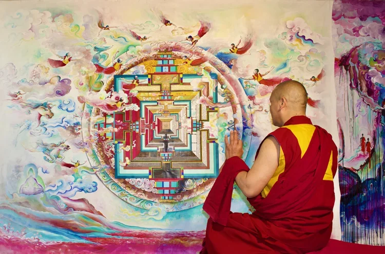 A Buddhist monk in traditional red and yellow robes sitting cross-legged and meditating in front of a colorful wall painting of a mandala with flying figures, clouds, and a vibrant background.