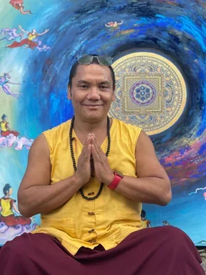 Lama Tashi sitting cross-legged in front of a vibrant, colorful mural with spiritual and abstract imagery, holding his hands in a prayer gesture, wearing a yellow sleeveless shirt, sunglasses on his head, and a beaded necklace.
