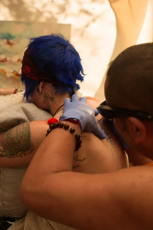 A person with blue hair getting a tattoo on their shoulder from a tattoo artist.