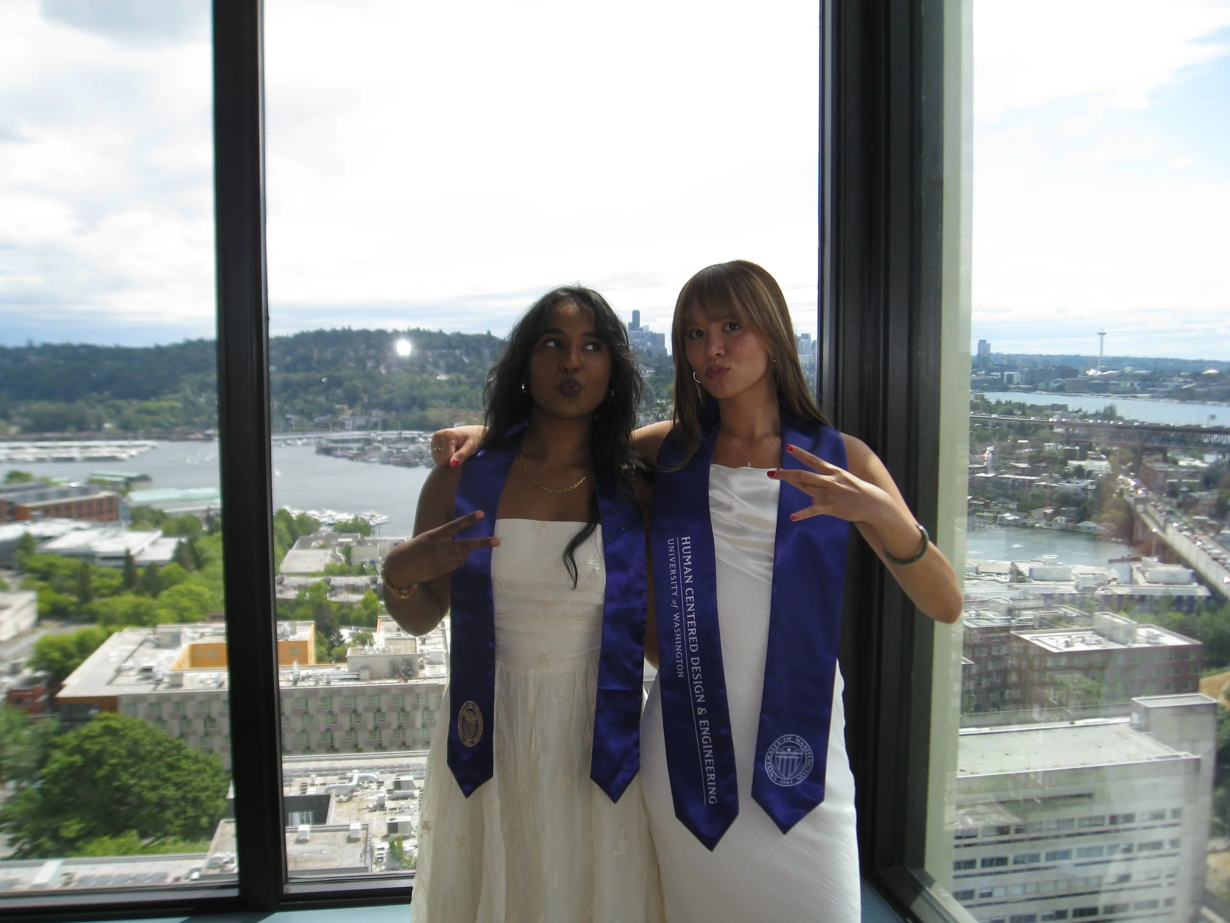 Two women, one Black and one Asian, standing together in front of a large window with a city and river view behind them. They are wearing blue sashes, and the woman on the left is wearing a white dress, while the woman on the right is wearing a white top. Both women are making a peace sign with their hands and have their arms around each other.