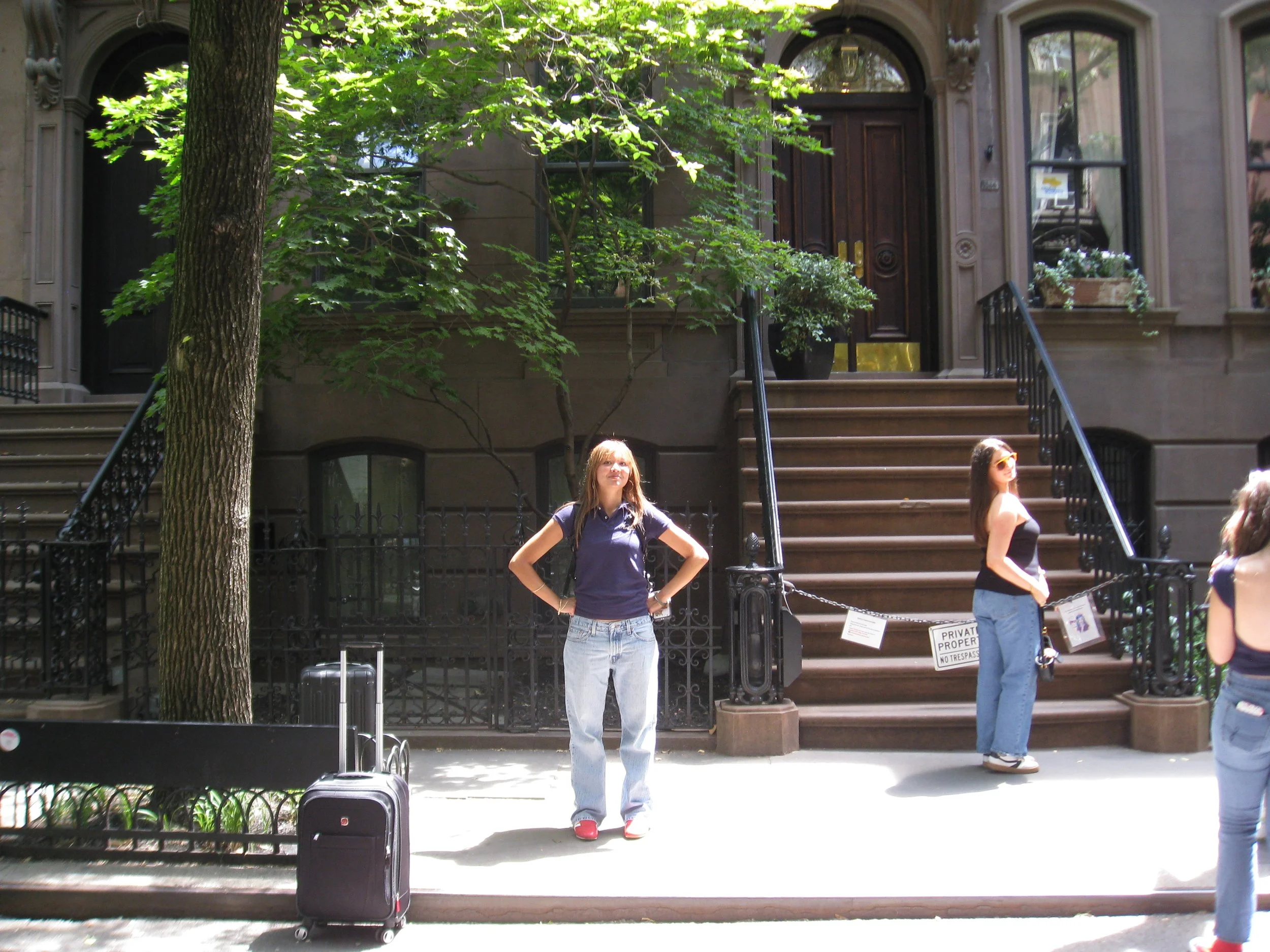 People standing and walking on the sidewalk in front of brownstone row houses with stairs, black railings, and large windows; a tree with green leaves partially blocks the view.
