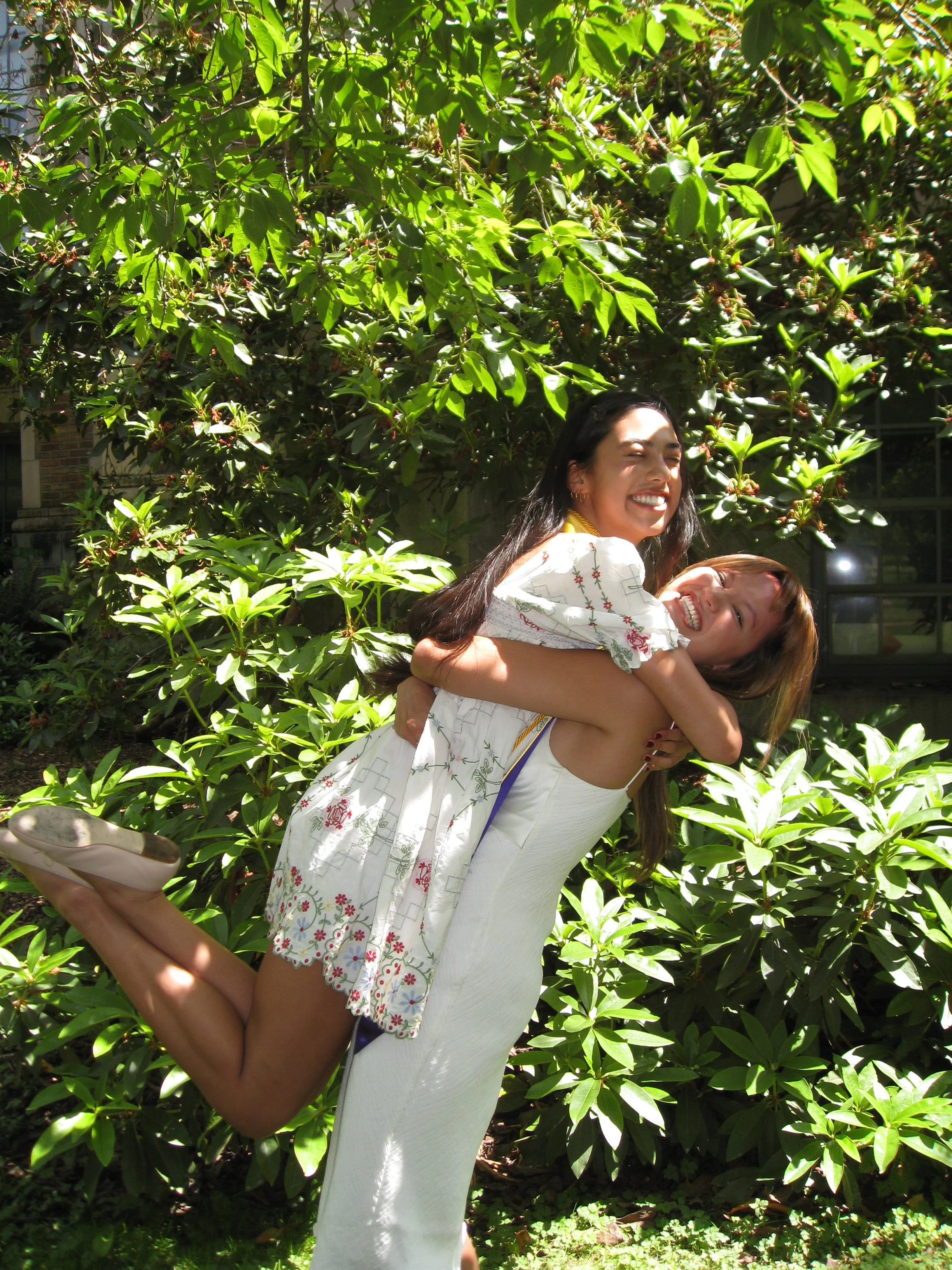 Two women smiling and posing playfully outdoors in front of lush green foliage, with one woman standing and the other being carried on her back.