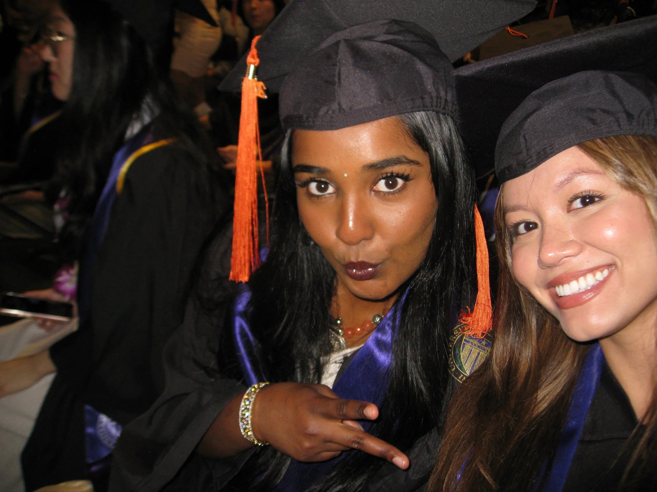 Two women at a graduation ceremony wearing caps and gowns, one with dark hair and makeup, and the other with lighter hair and a big smile, standing closely together.
