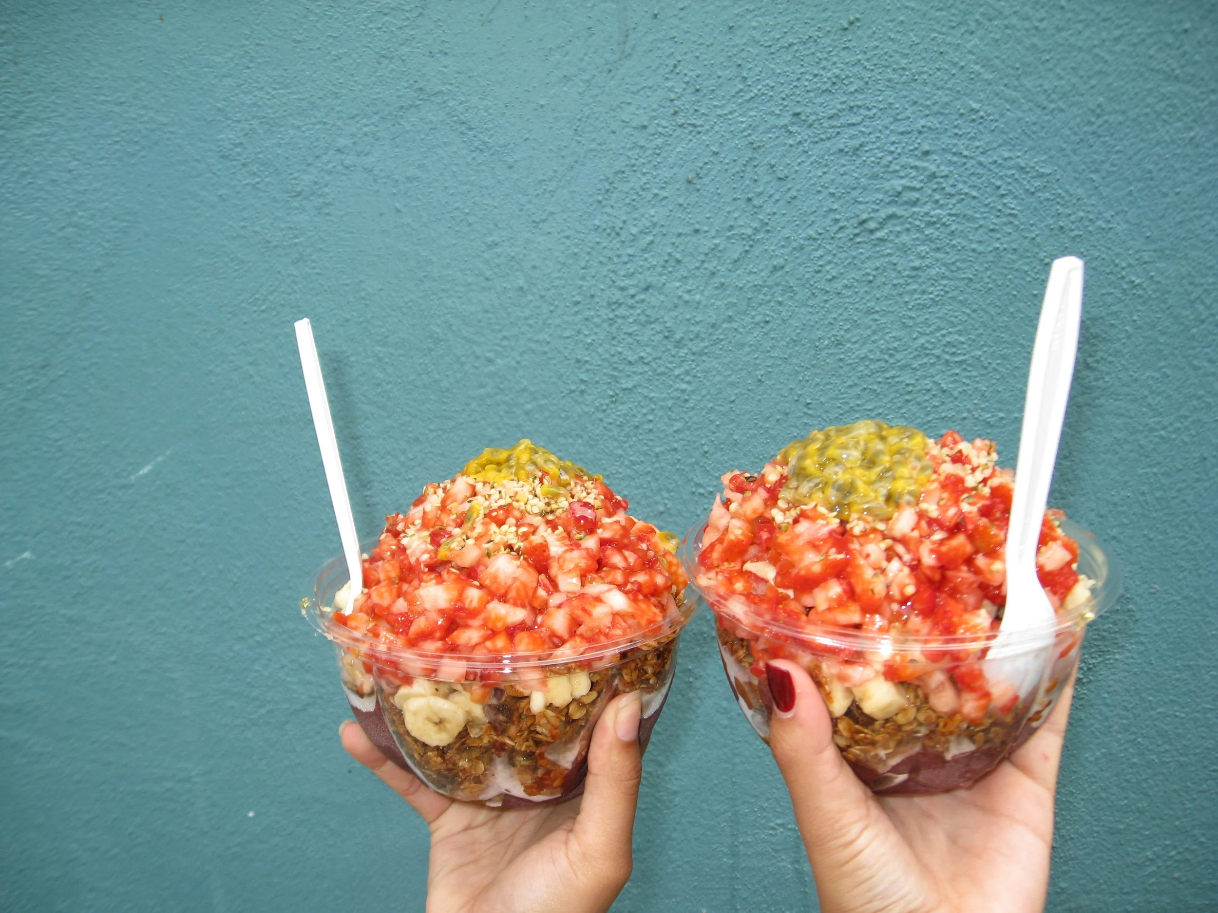 Two cups of colorful rice pudding topped with fruit, nuts, and syrups, held by hands against a teal wall background.