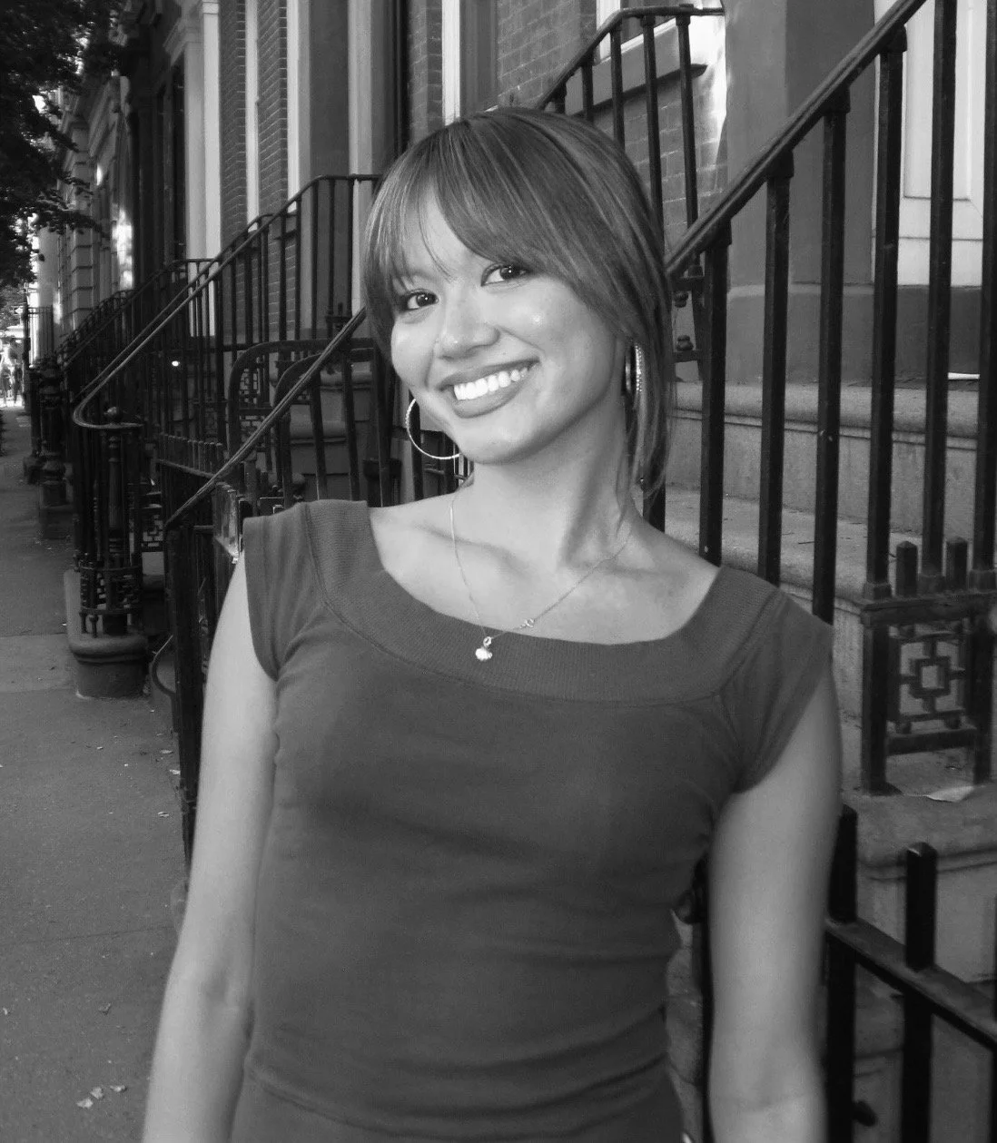 A woman with medium-length hair and a bright smile, wearing hoop earrings and a necklace, standing on a city sidewalk with brick buildings and stairs in the background.