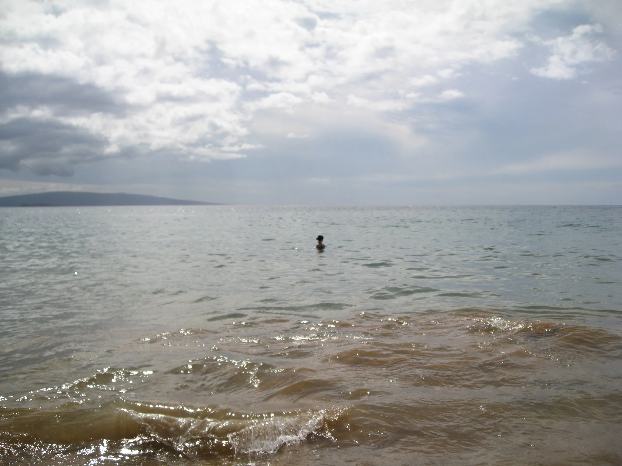 Overcast sky with clouds reflected on calm ocean waters, a person wading in the water near the shore, distant landmass in the background.