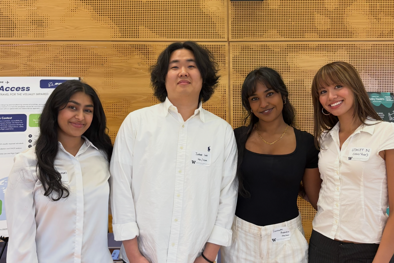 Four diverse young adults standing together indoors in front of a wooden wall, smiling at the camera, two women wearing white shirts with name tags, background includes informational posters.