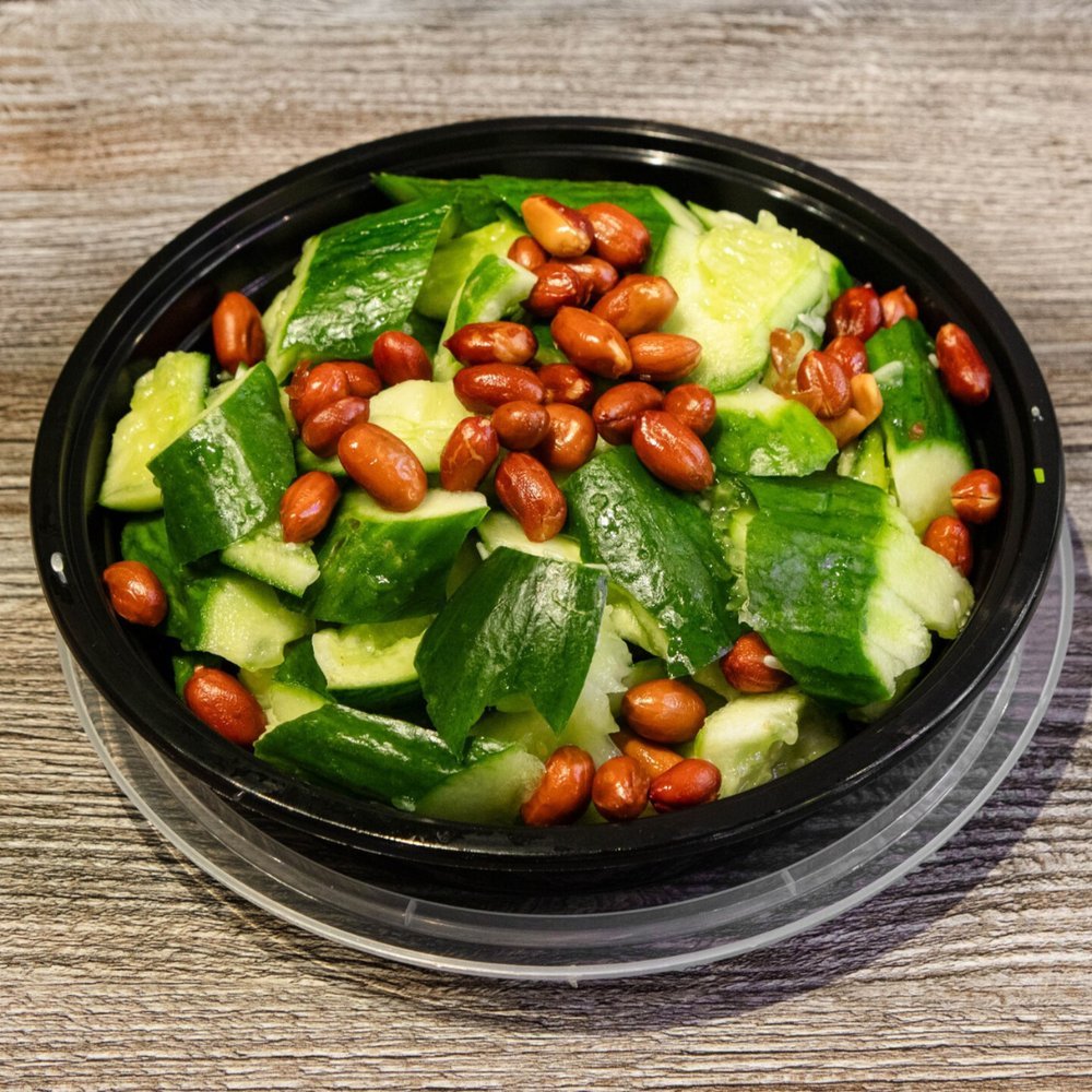 Fresh cucumber slices topped with toasted pine nuts in a black plastic salad container on a wooden surface.