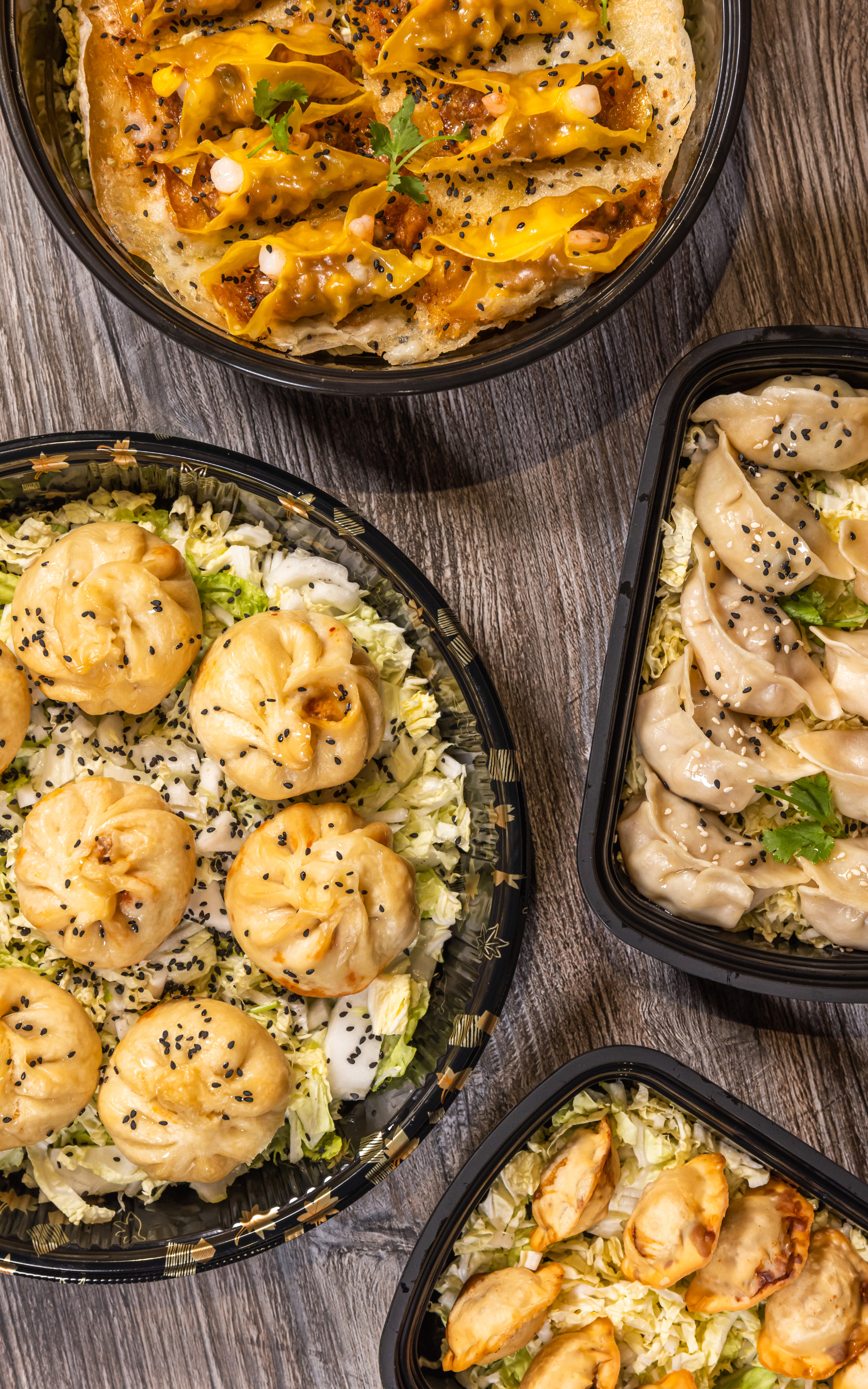 Various types of Asian-style dumplings and siu mai with black sesame seeds on top, served in black plastic takeout containers with shredded lettuce and cabbage on a wooden surface.