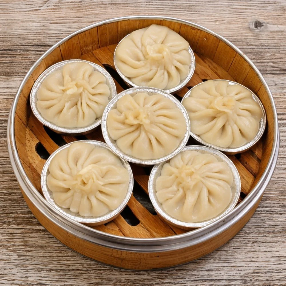 Six steamed dumplings with pleated tops in aluminum cups inside a round bamboo steamer basket on a wooden surface.