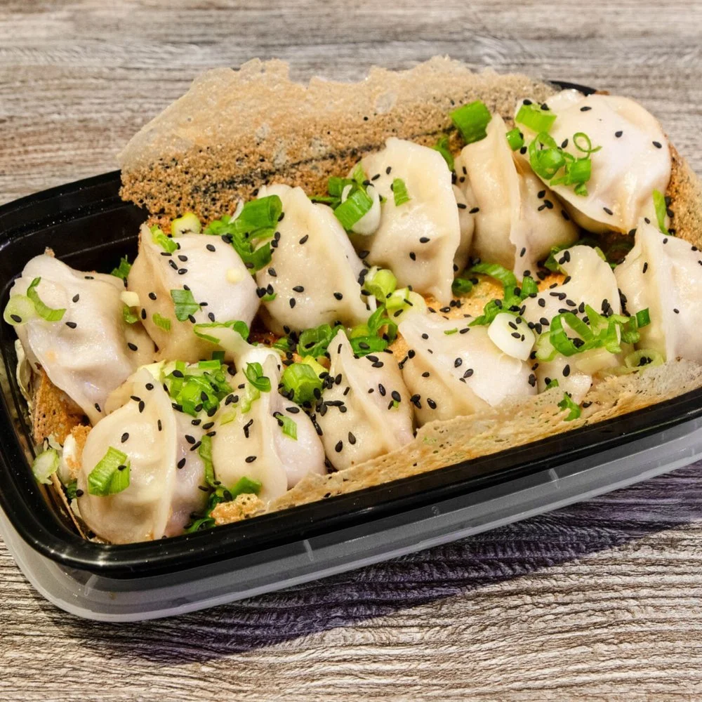 A black rectangular tray of dumplings garnished with chopped green onions and black sesame seeds, placed on a wooden surface.