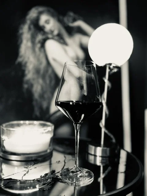 A wine glass filled with red wine in foreground, a lit candle, a decorative lamp, and a blurred black and white photo of a woman with long hair in the background.