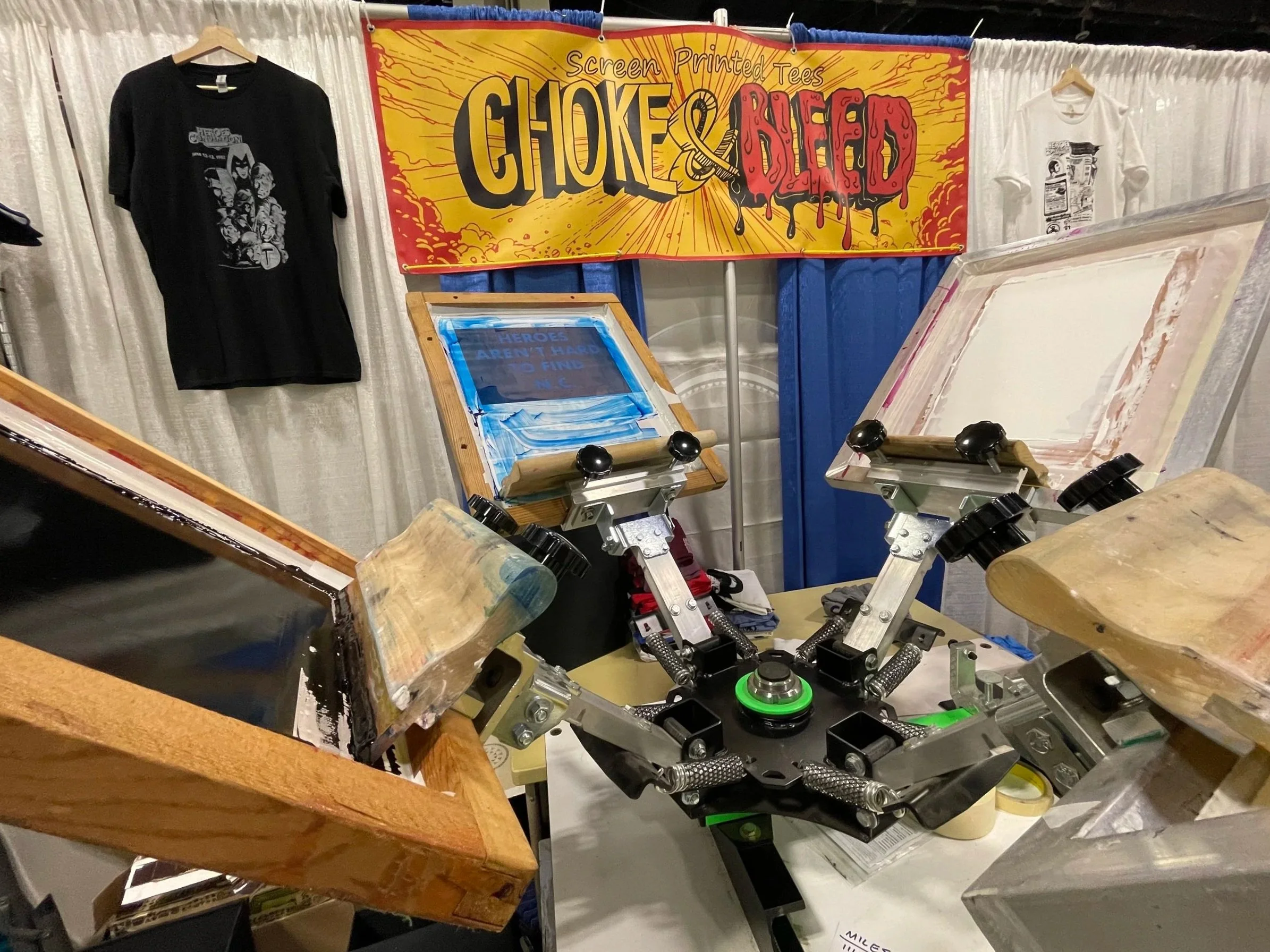 A booth displaying screen-printed t-shirts with a banner that reads 'Screen Printed Tees' in a comic book style. The booth has a screen printing setup with wooden frames, ink, and printing equipment, alongside t-shirts hanging on the wall.