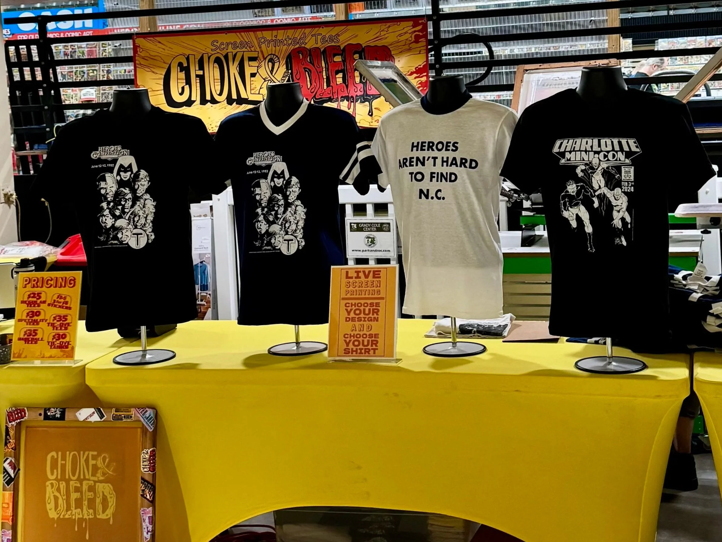 Display of T-shirts with superhero and convention themes at a booth, with signs indicating prices and customization options, and a brightly colored sign that reads 'Choke & Bled' in the background.