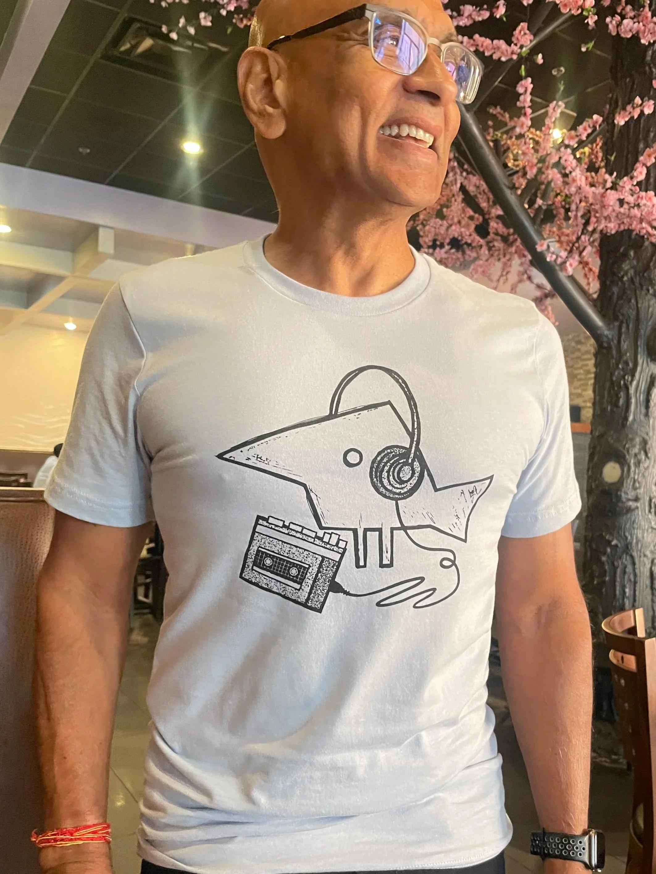 Close-up of a man wearing glasses and a white T-shirt with a graphic design of a cassette tape, headphones, and a microphone. He is smiling, with pink cherry blossoms in the background, inside a restaurant or cafe.