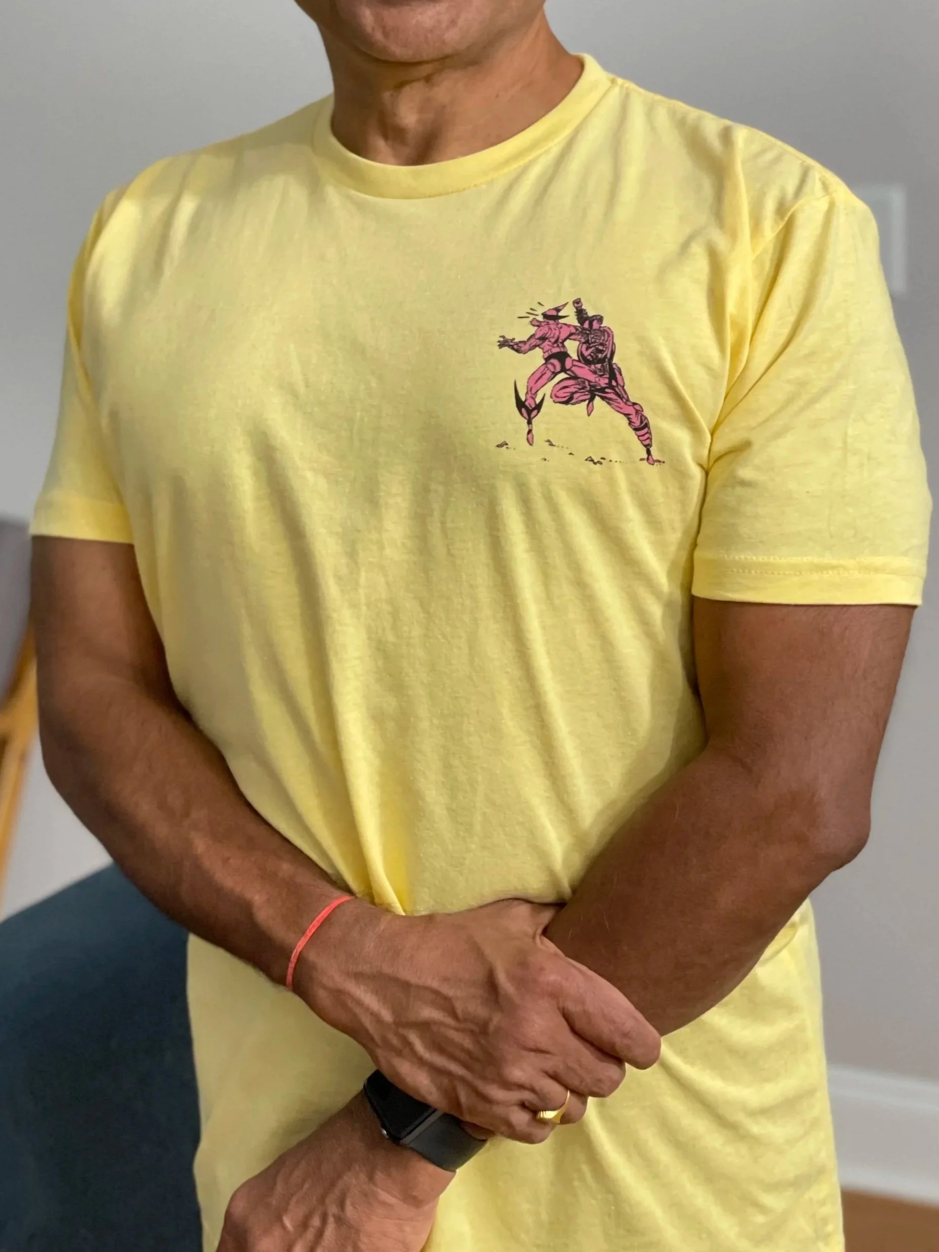 Close-up of a person wearing a yellow T-shirt with a graphic of a fighting characters on the front. The person's arm is crossed and holding their hand, showing a wristwatch and a ring on the finger.