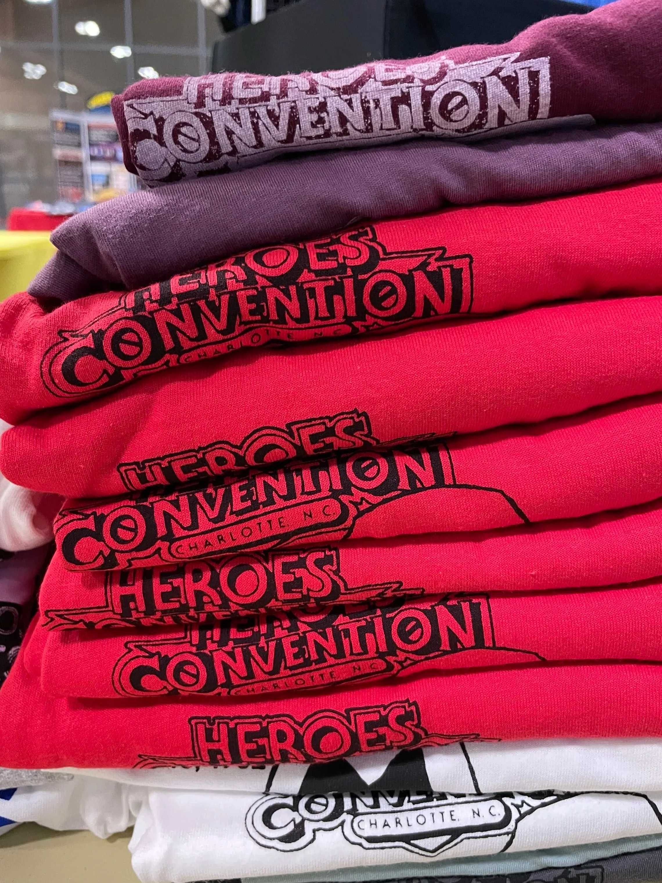 Stacked folded t-shirts with 'Heroes of Konvention Charlotte, NC' printed in black on red, pink, purple, and white shirts.