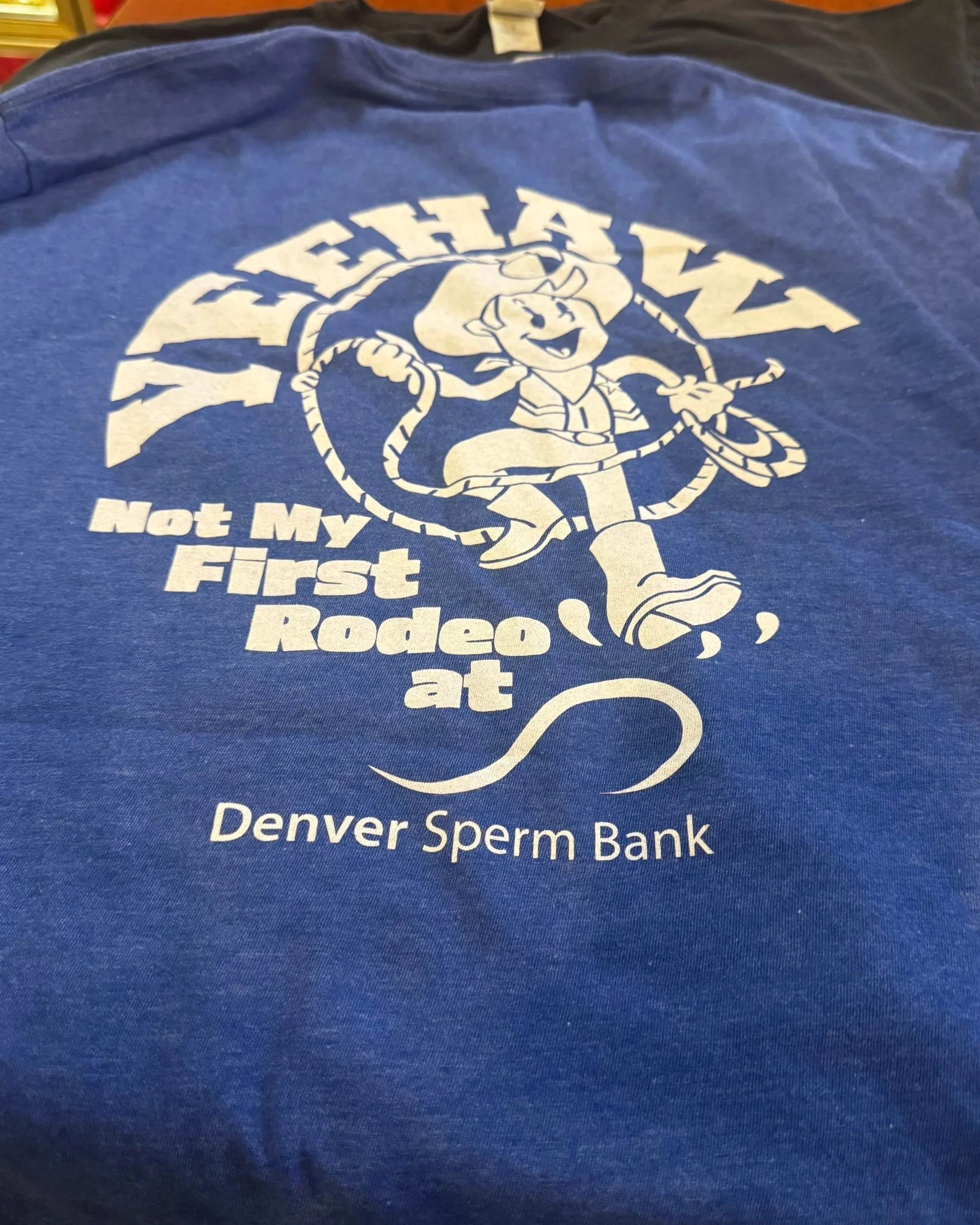 Blue t-shirt or sweatshirt with white printed graphic of a cartoon pig dressed as a cowboy, holding a lasso. Text on the shirt reads: "Not My First Rodeo," and at the bottom, "Denver Sperm Bank."