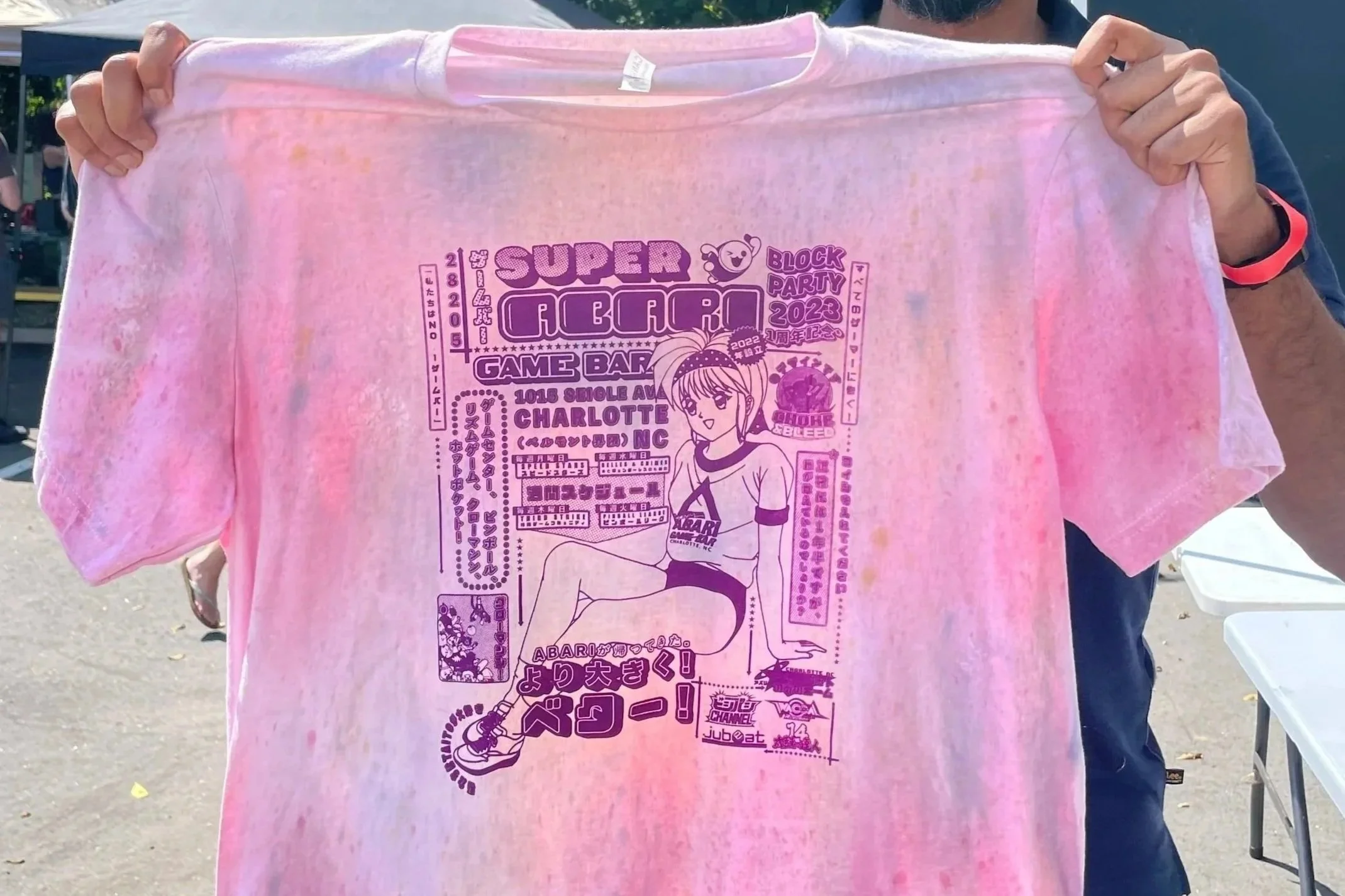 Person holding up a pink T-shirt with a purple and black graphic design for the Super Agraph 2023 block party, featuring anime-style artwork and text.