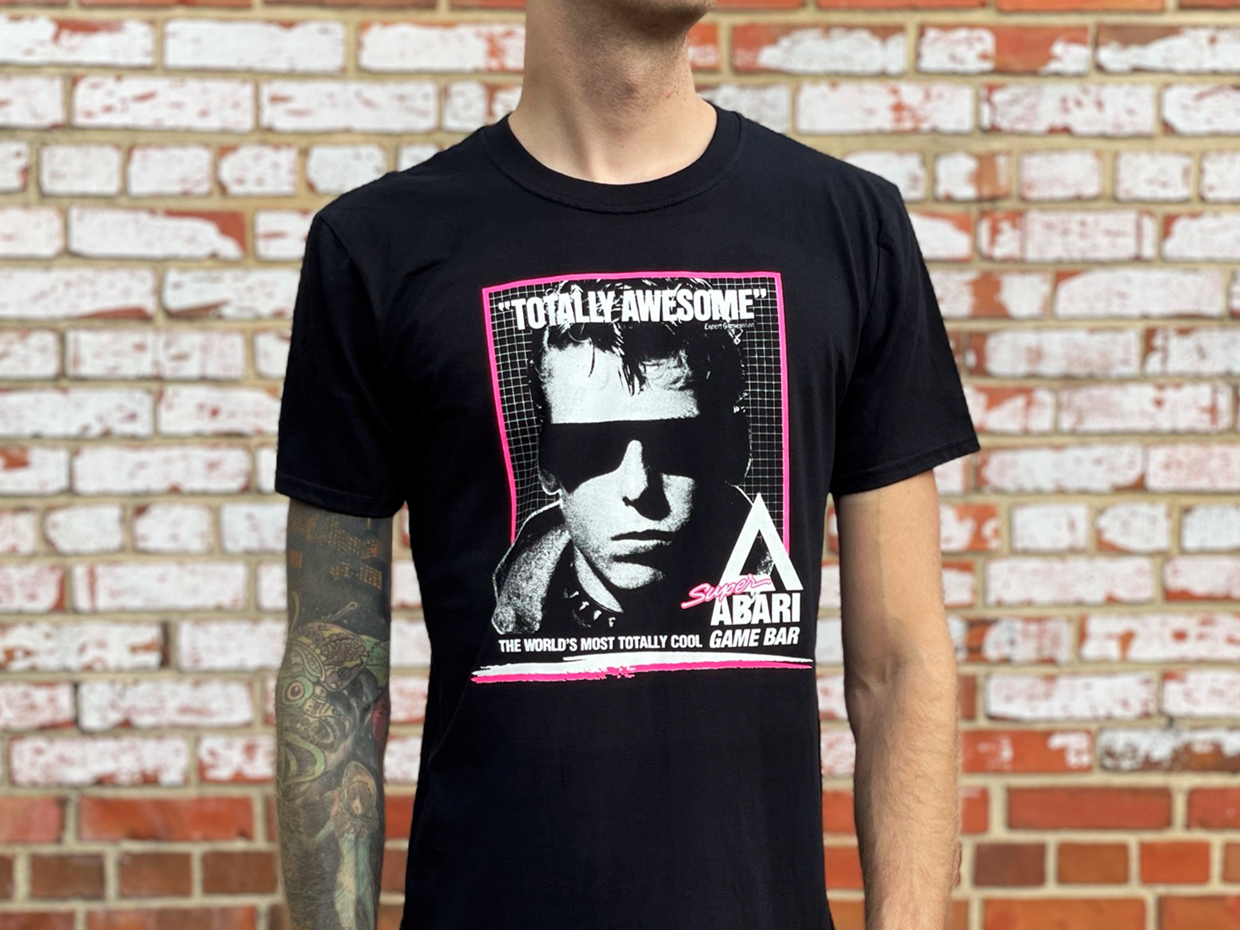 Close-up of a person wearing a black T-shirt with a graphic design, standing in front of a brick wall. The T-shirt features a black and white portrait of a man with sunglasses, text that reads "TOTALLY AWESOME" and "THE WORLD'S MOST TOTALLY COOL GAME