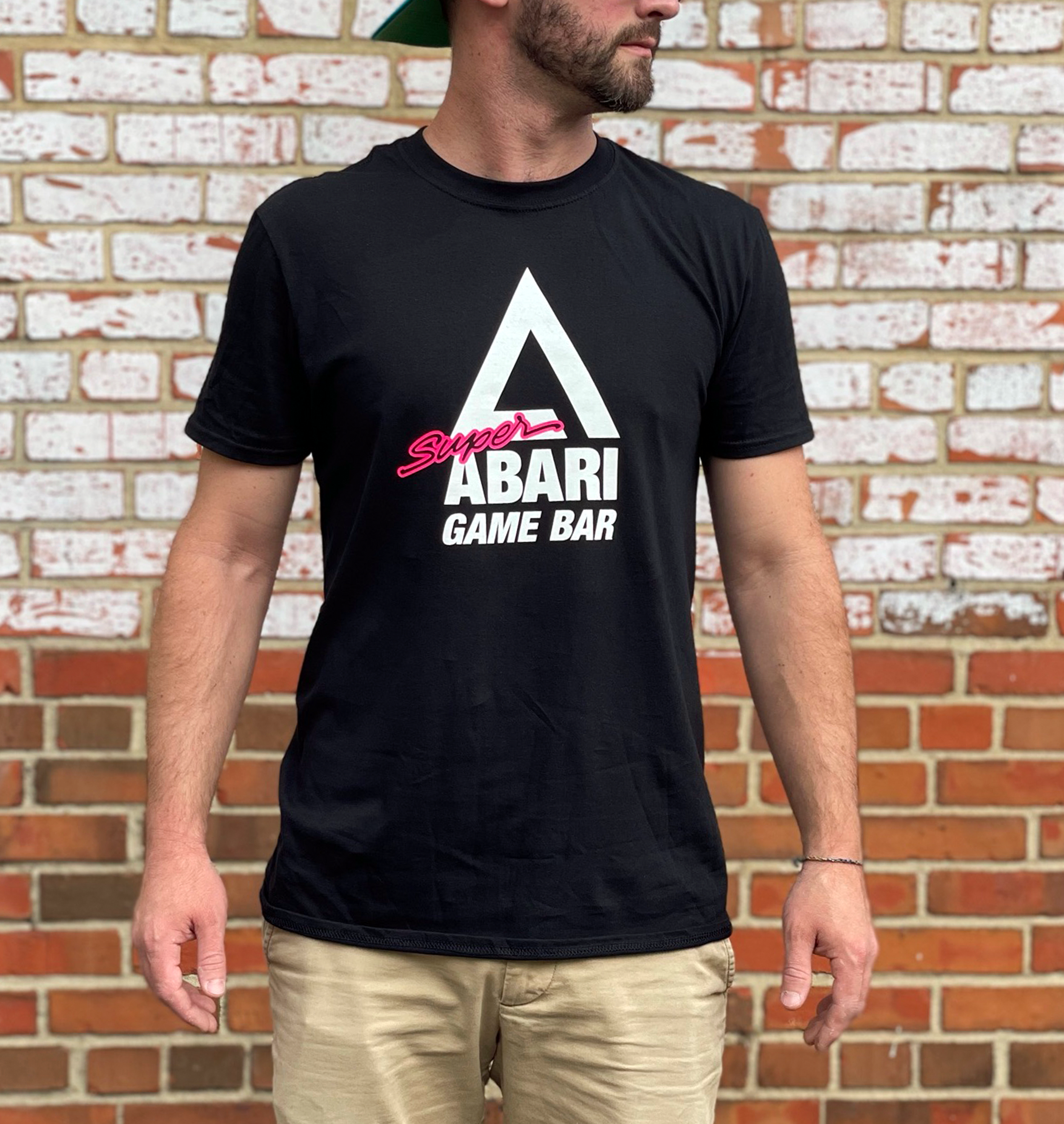 A man wearing a black T-shirt with the text 'Super ABARI GAME BAR' in front of a brick wall, with only part of his face visible, slightly turned to the right, and arms hanging down with hands relaxed.