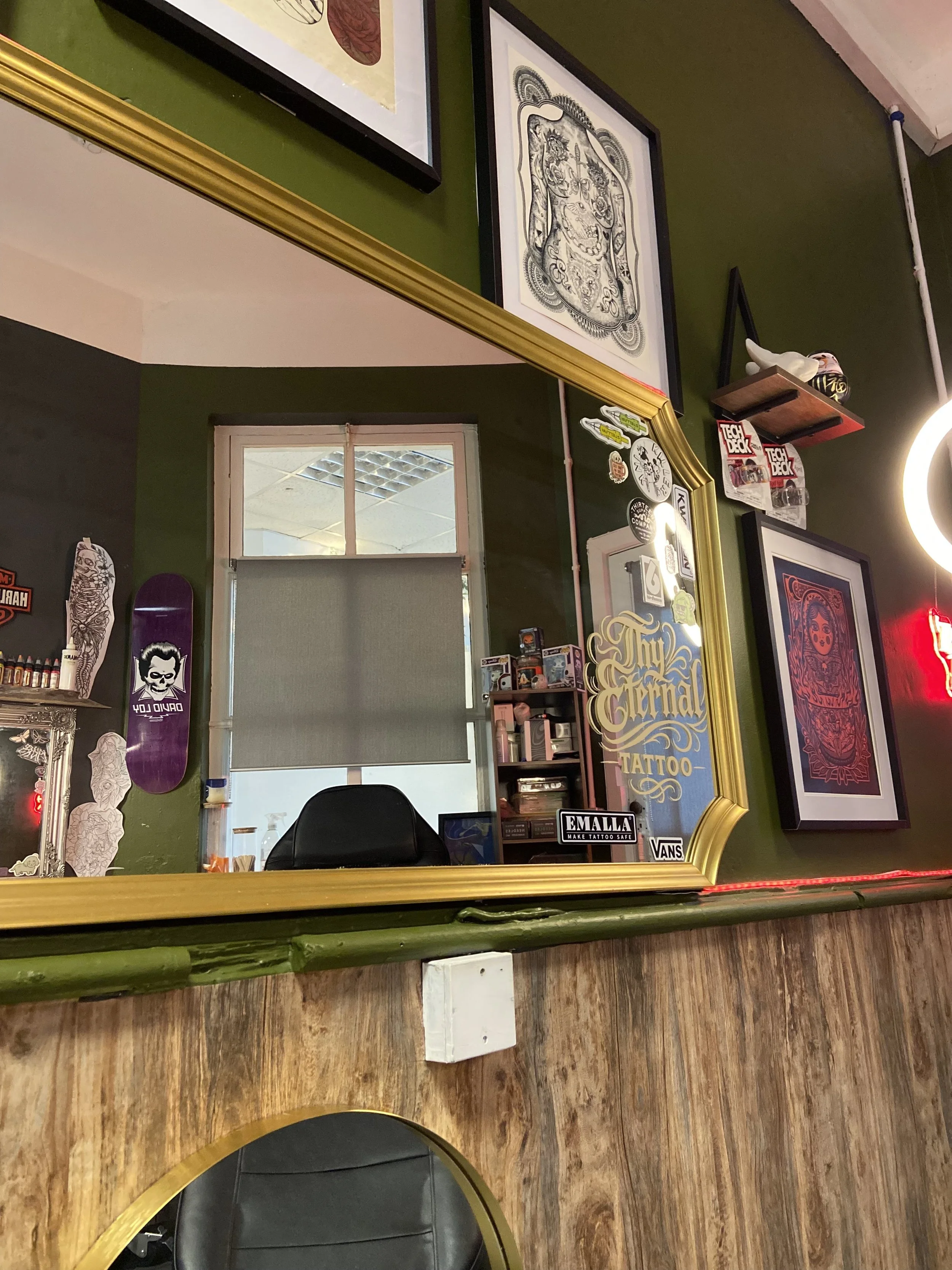 A wall mirror in a tattoo shop reflecting framed artwork, stickers, and a window with a roller shade. The wall is painted green with various tattoo-related decorations and posters. A chair with a gold rim is visible at the bottom of the image.