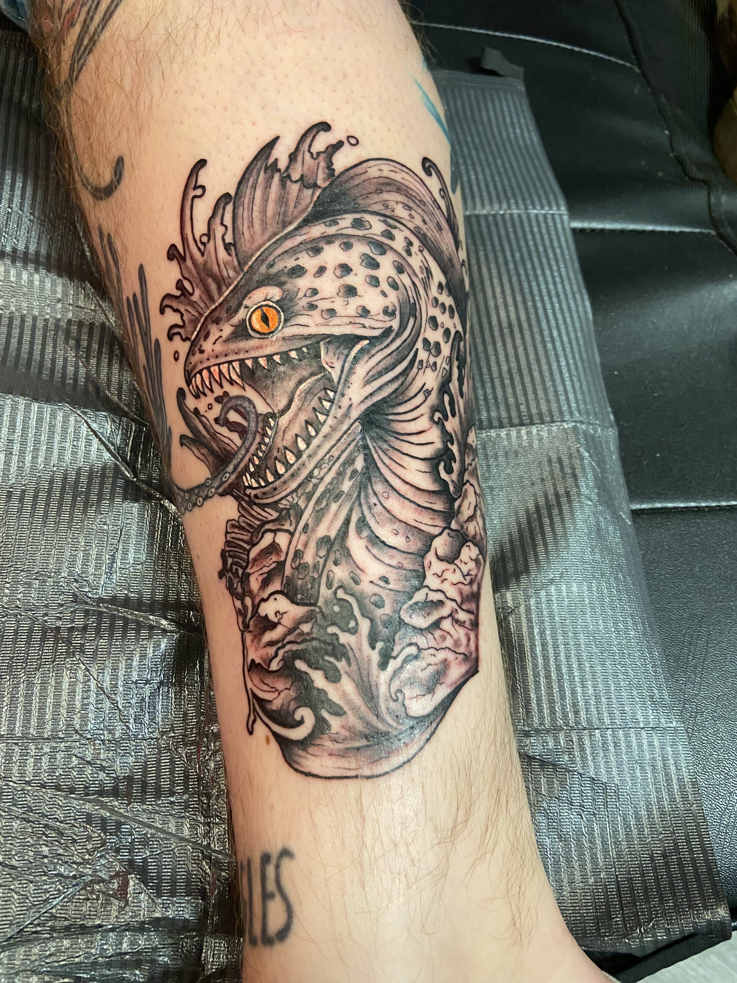 A black and gray tattoo of a fierce-looking dinosaur with orange eyes on a person's arm, with swirling elements and clouds at the bottom.