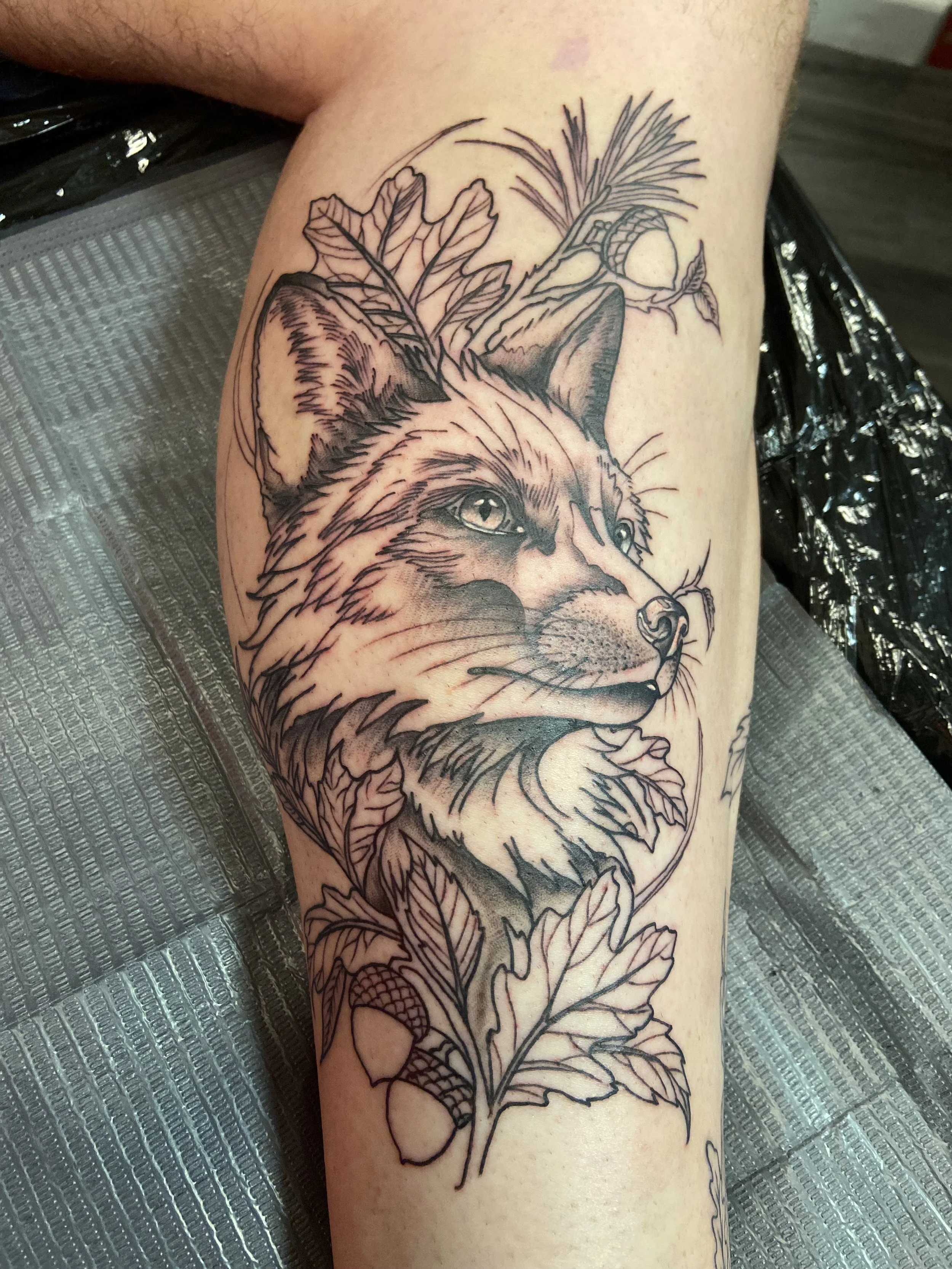 Black outline tattoo of a wolf's face with leaves, pinecones, and pine needles around it on a person's thigh.