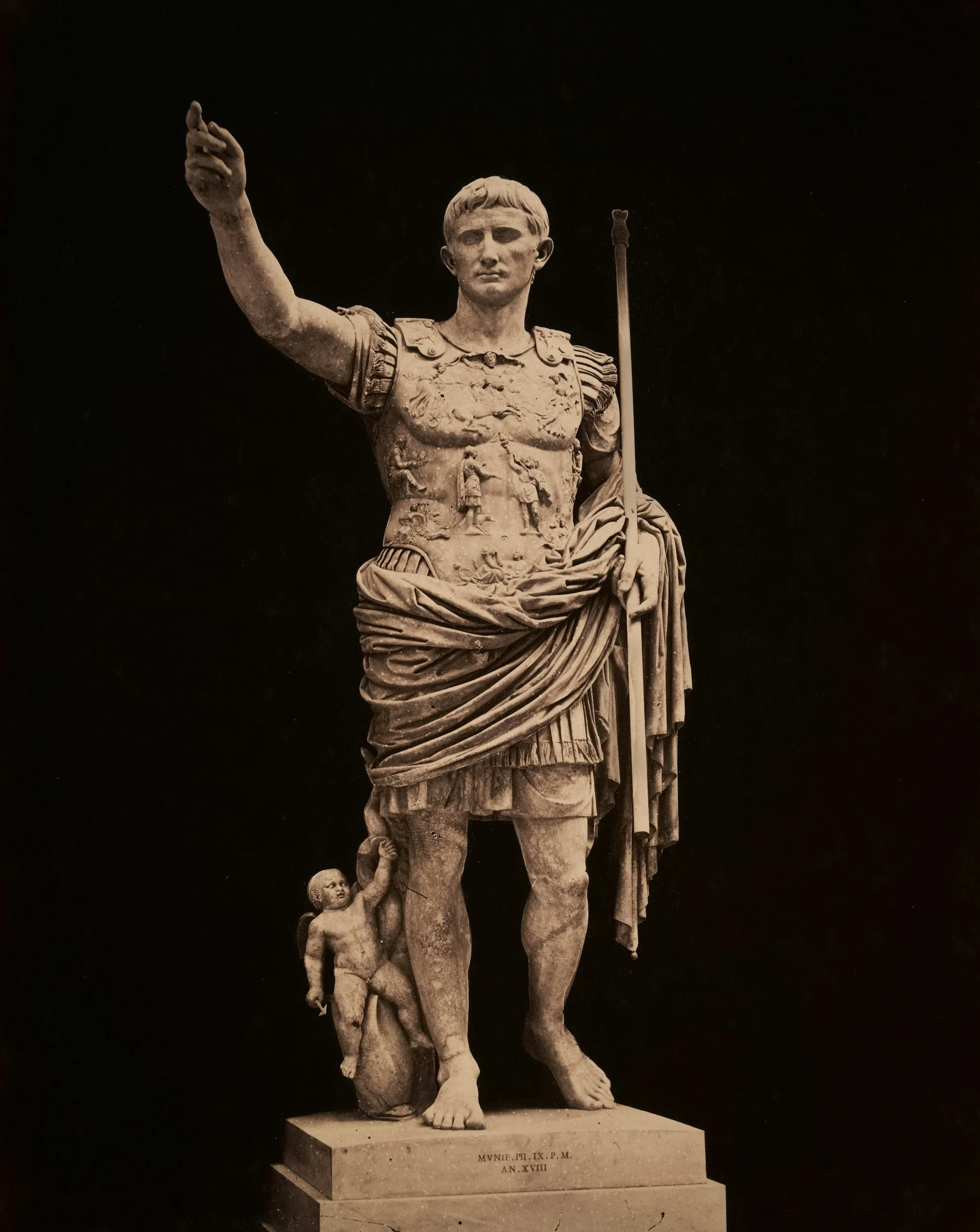 A marble statue of a Roman soldier or emperor holding a staff in his left hand, with a raised right arm. The statue features detailed armor and a cloak draped over his shoulder. There is a small child or cherub holding onto his leg at the base of the statue.